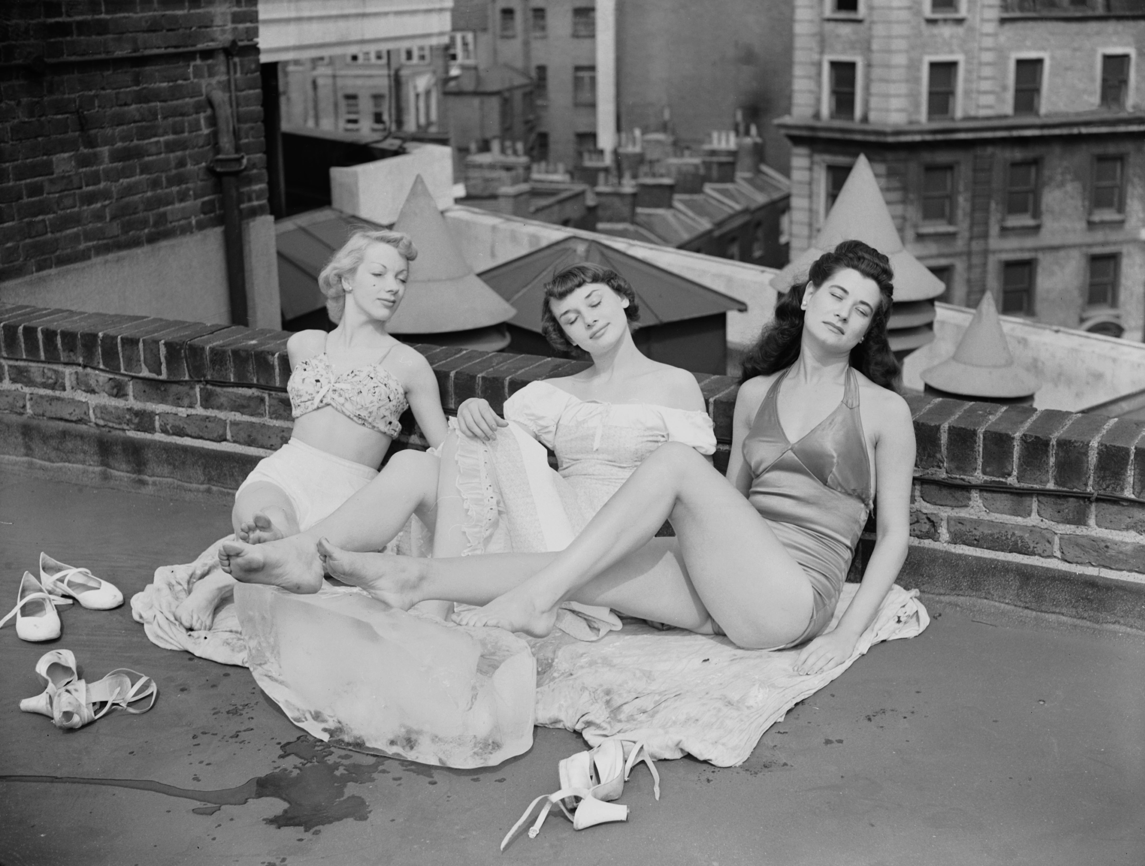 Three women in 1950s-style swimsuits relax on a rooftop, sitting on towels with an ice block, shoes set aside. Background features urban buildings