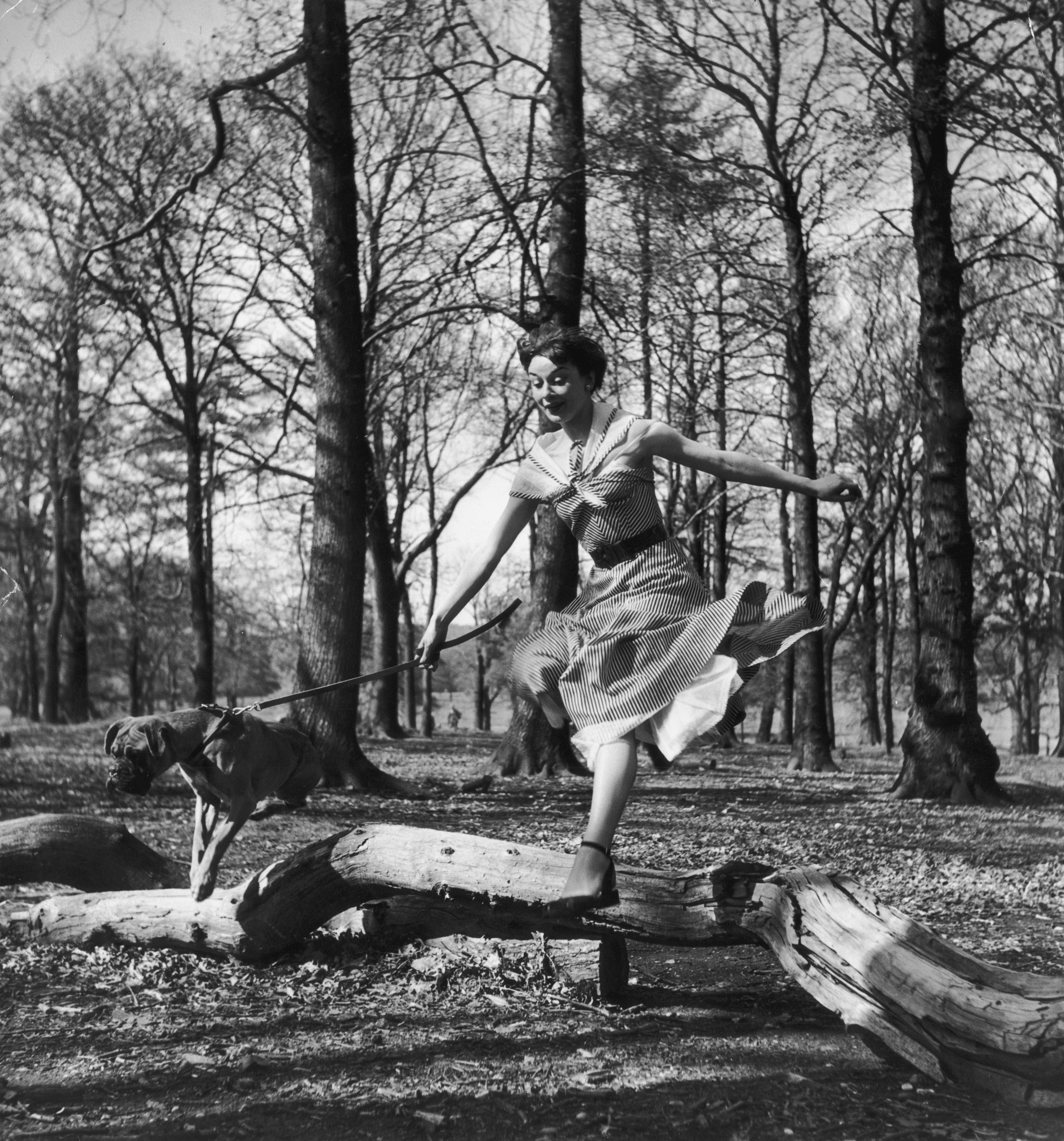 Audrey Hepburn joyfully jumps over a fallen tree, playfully handling a leashed dog in a forest setting
