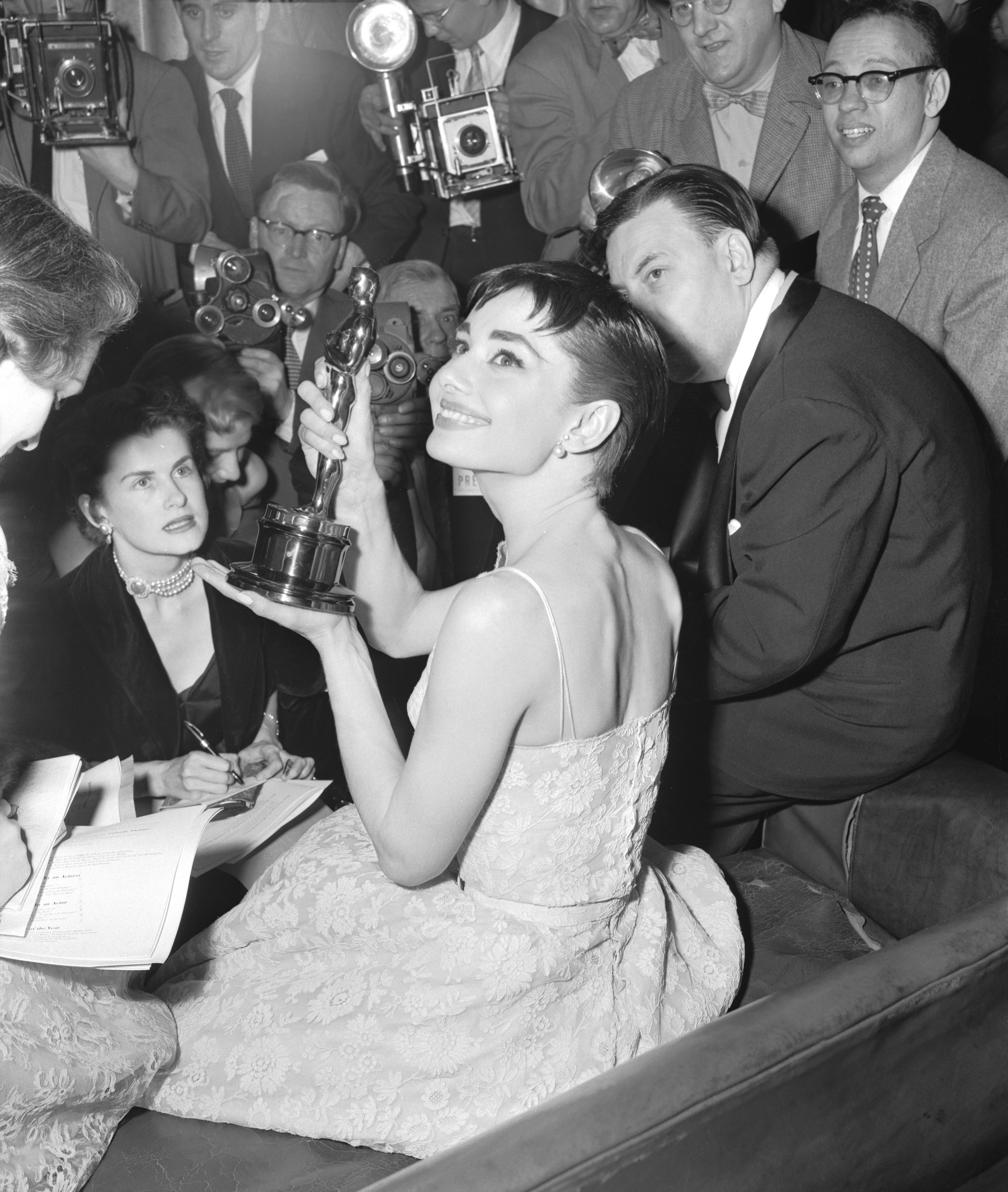 Audrey Hepburn in elegant lace dress holding an award, surrounded by photographers at a press event