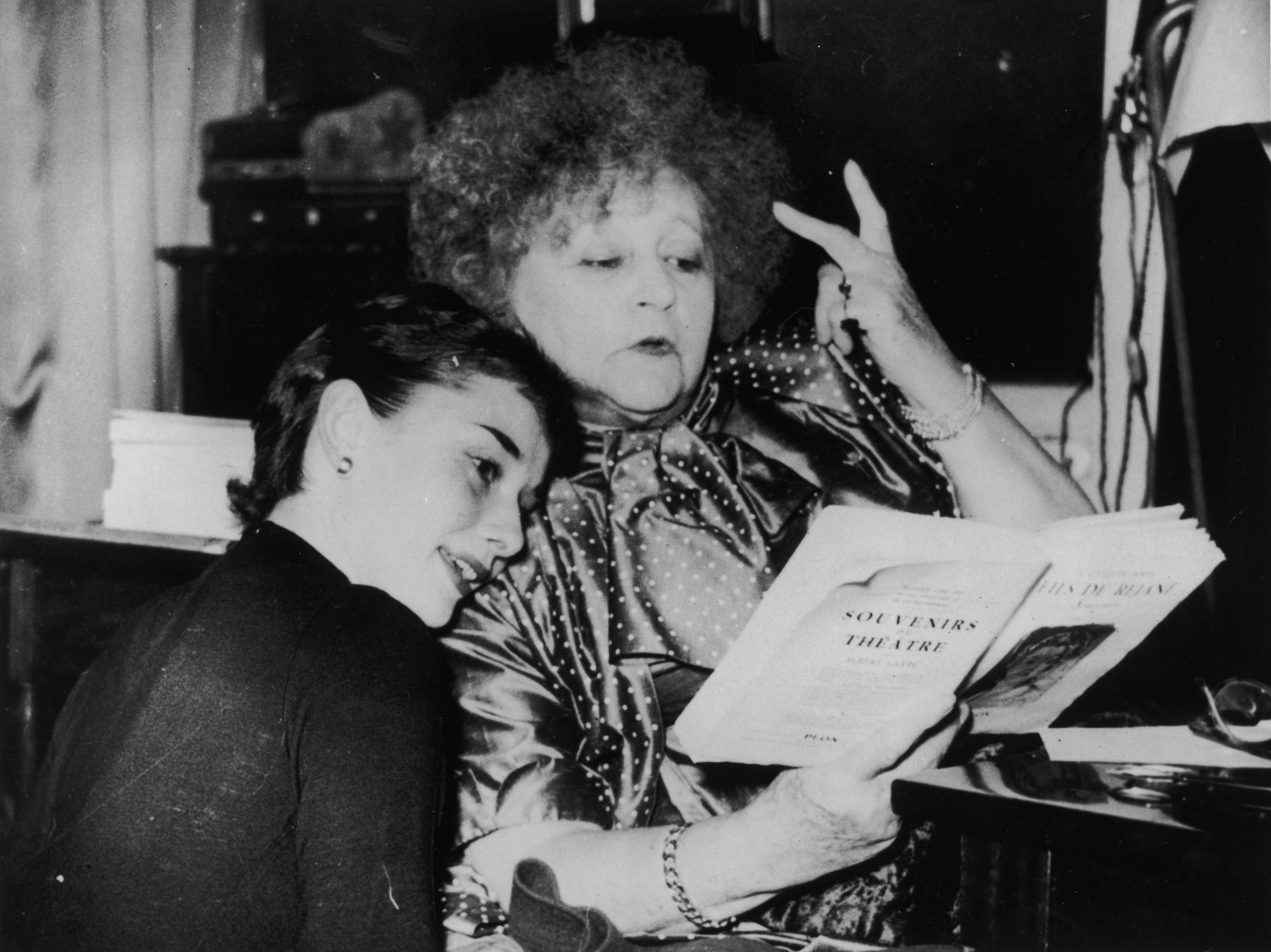 Colette reads a book aloud while Audrey leans on her shoulder, listening attentively. Both appear engaged in the activity