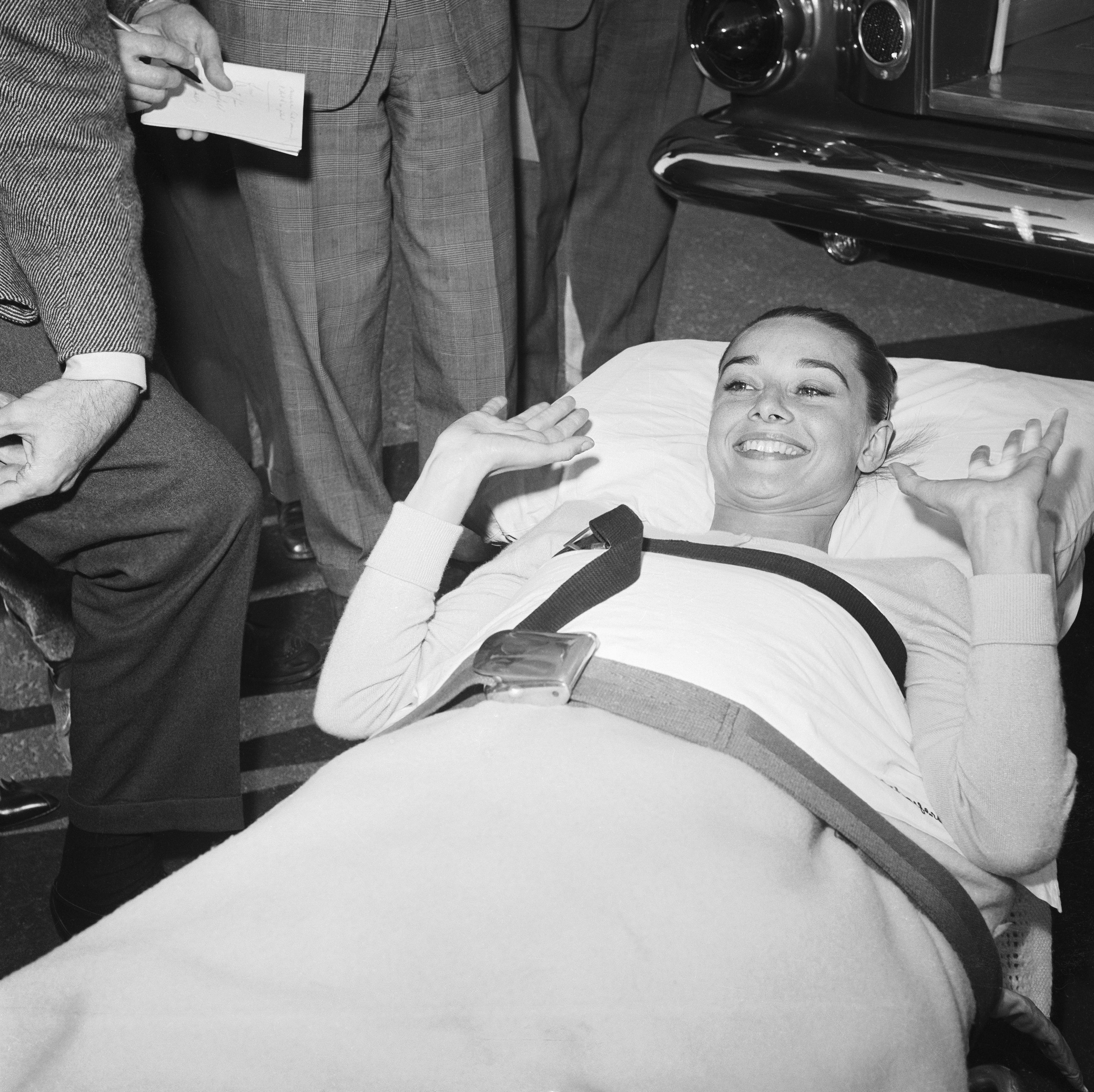 Audrey Hepburn lying on a stretcher with a seatbelt, smiling and gesturing, surrounded by people in formal attire