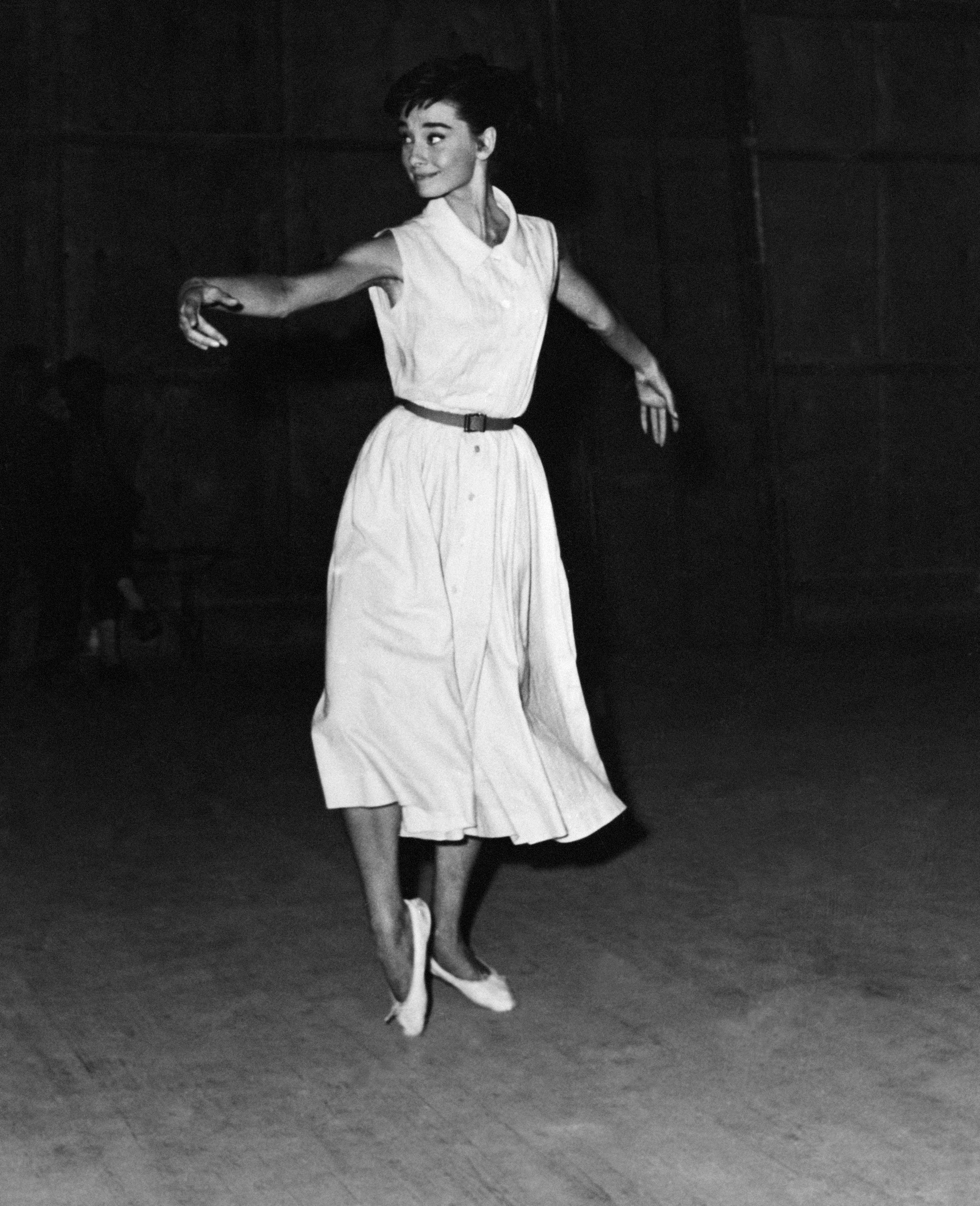 Audrey Hepburn dancing gracefully in a flowing dress, striking a poised, elegant pose in a dimly lit setting