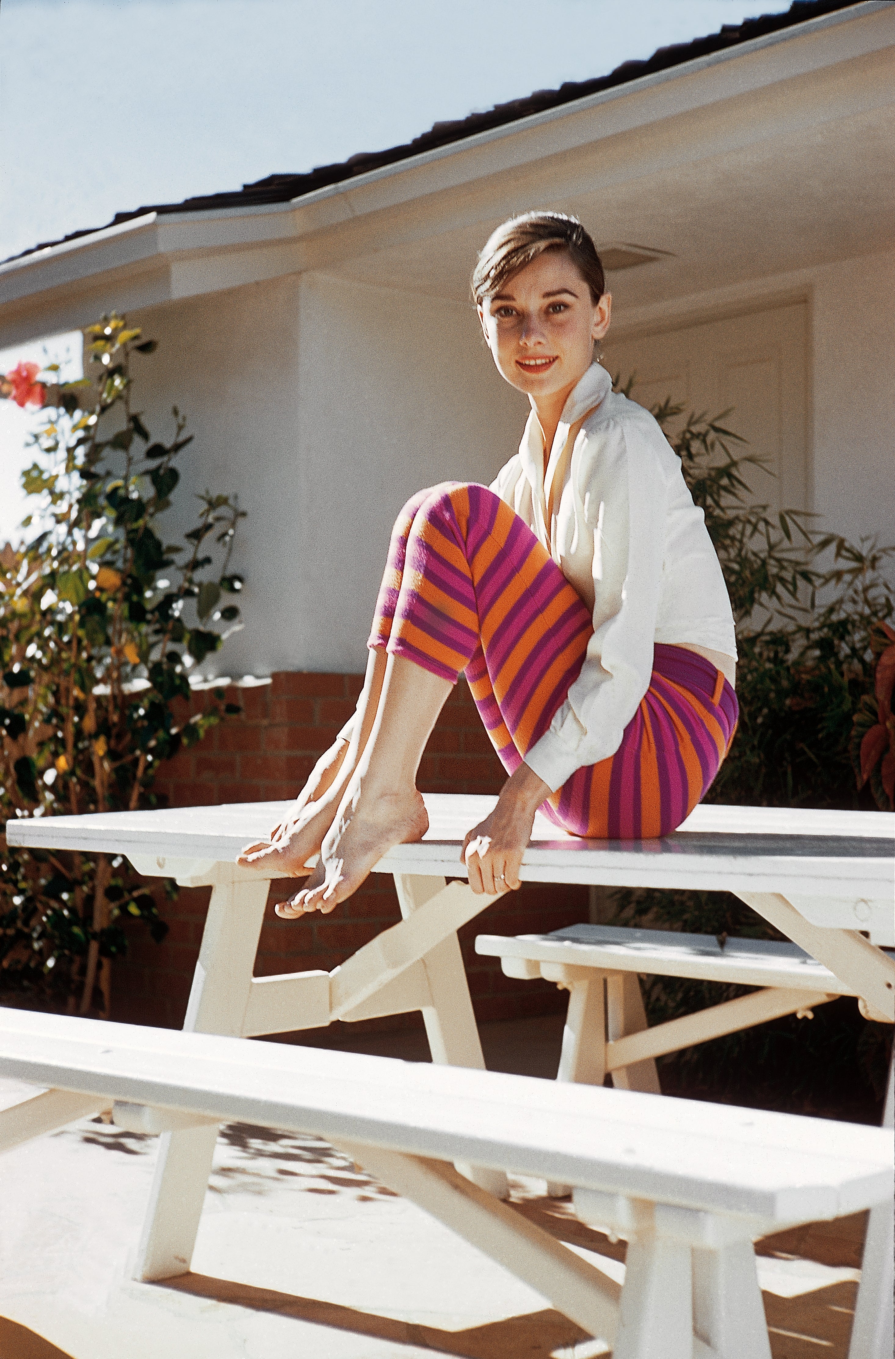 Audrey Hepburn sitting outdoors on a picnic table, smiling, wearing a white blouse and striped pants