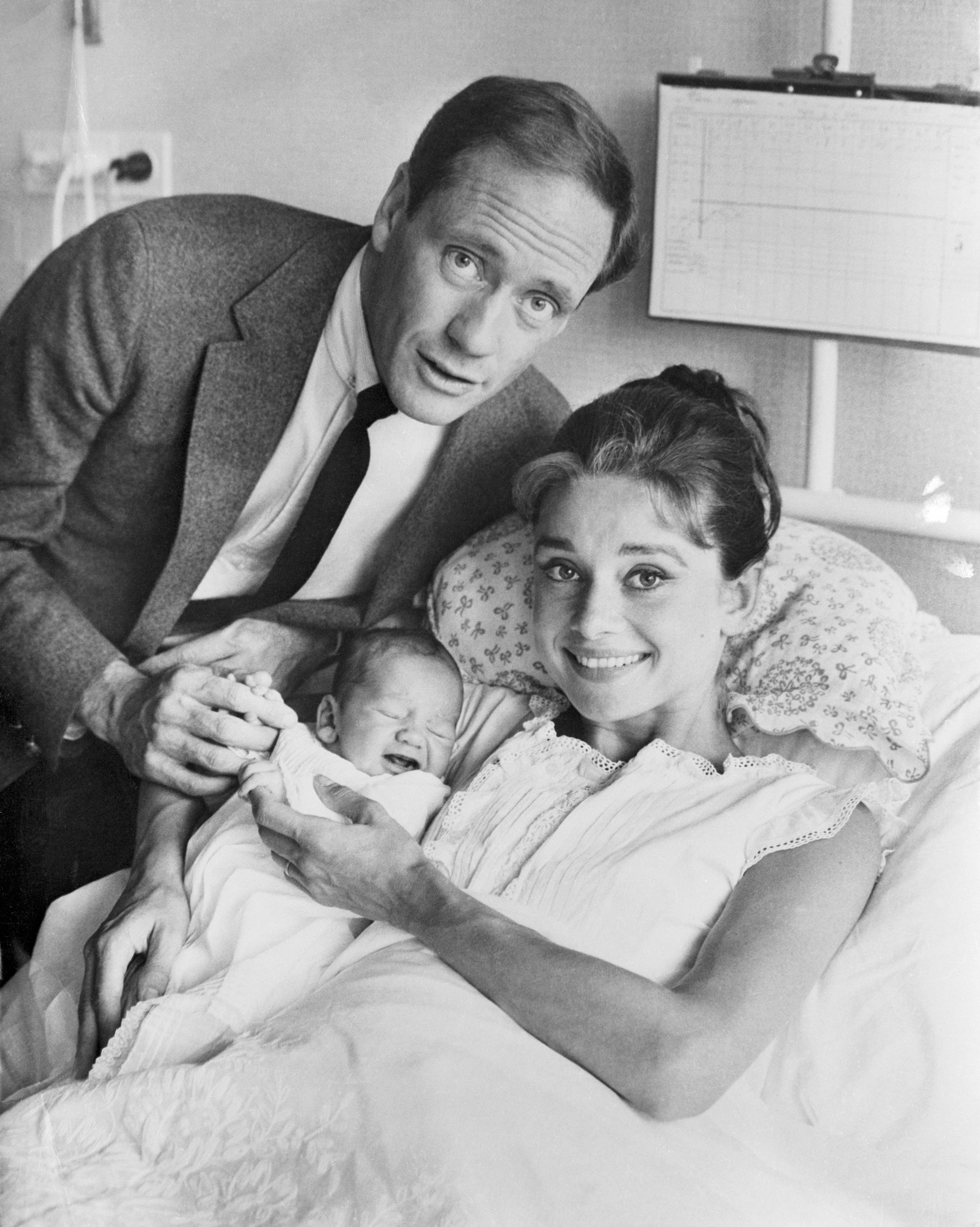 Audrey Hepburn in a hospital bed holds a newborn, smiling, with Mel in a suit standing beside them looking at the camera