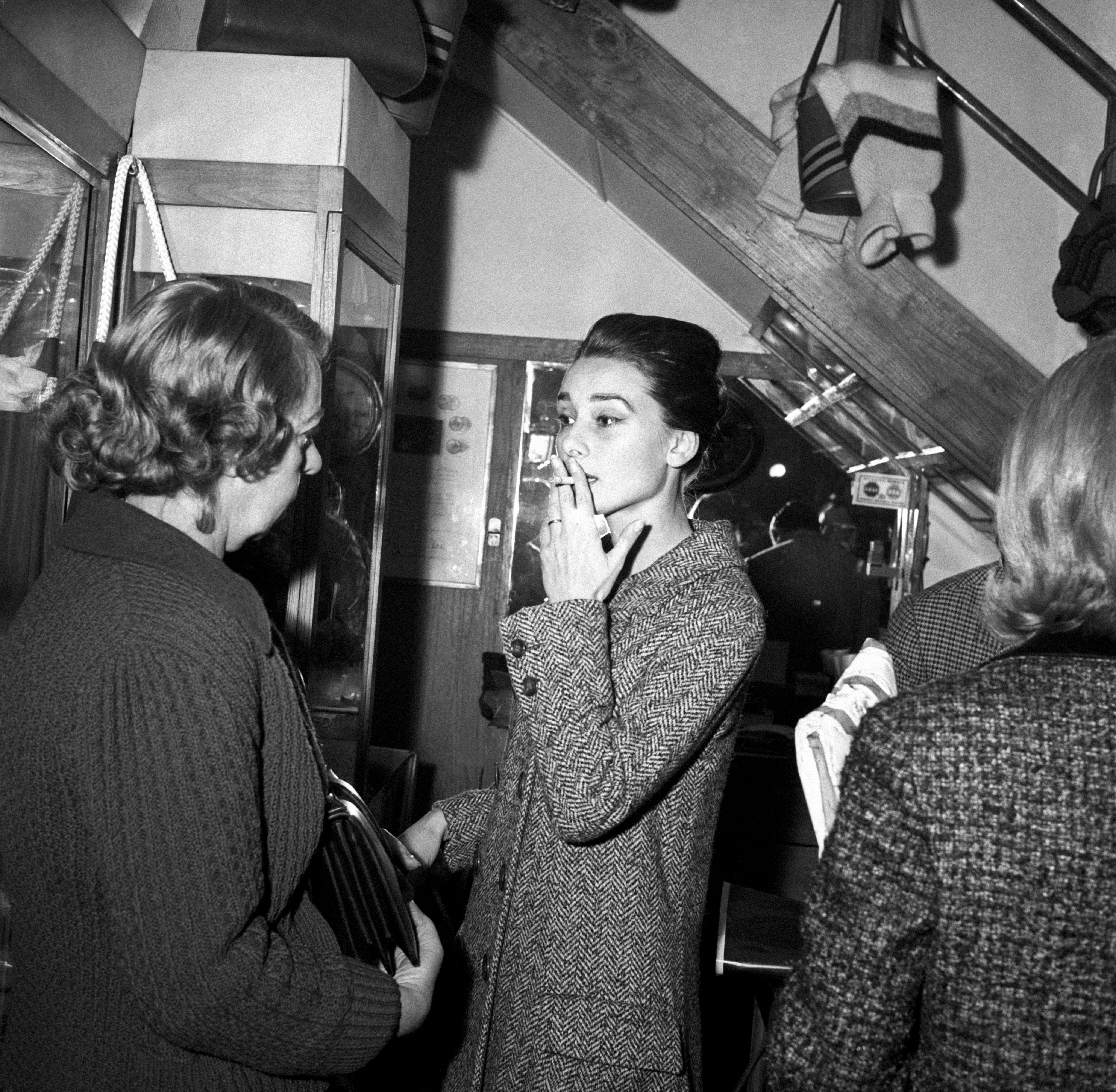 Audrey Hepburn in a chic, tailored coat smoking, engaging in conversation with others in a cozy, busy setting