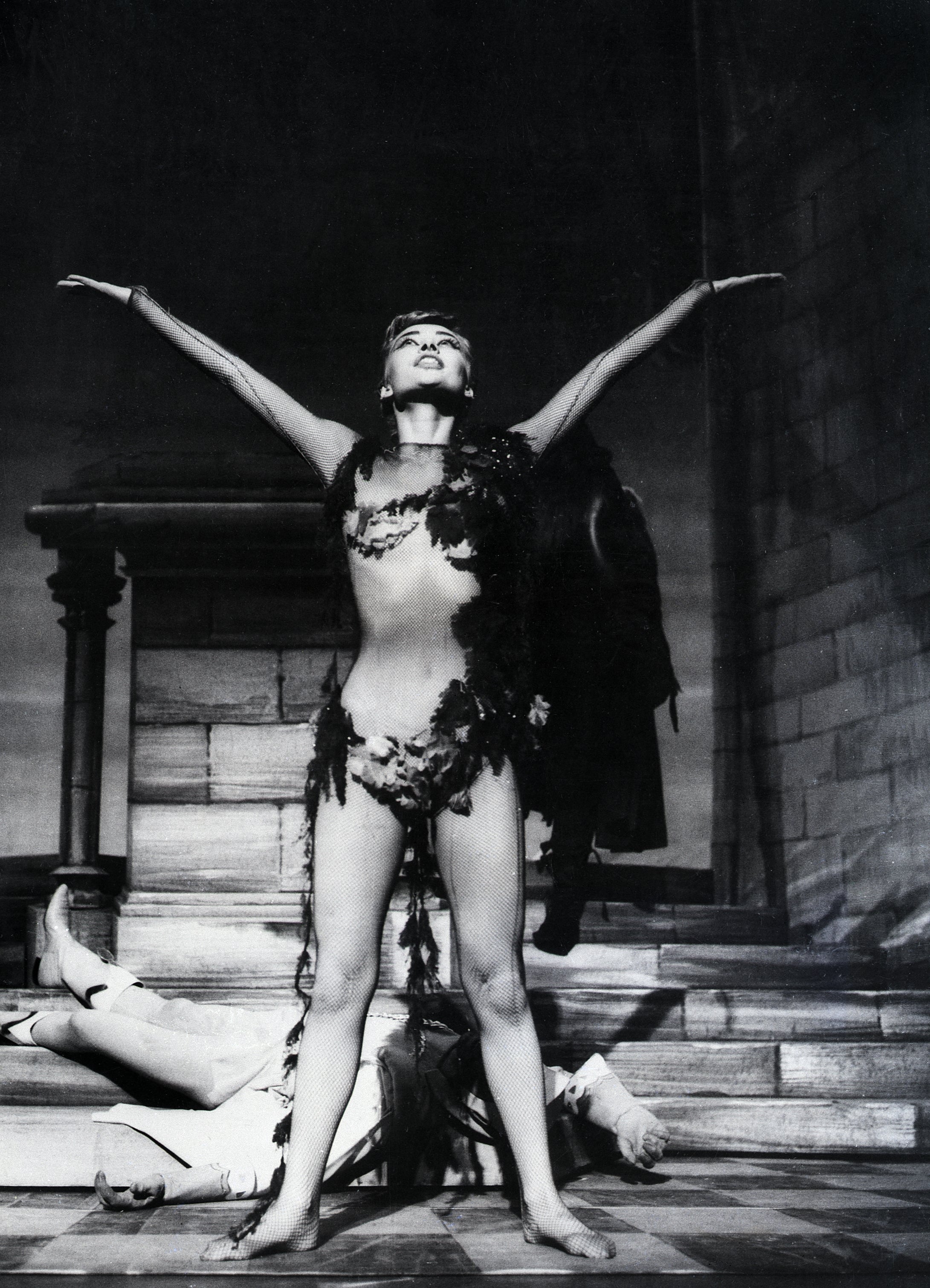 Audrey Hepburn on stage in theatrical costume, striking a dramatic pose with arms outstretched. Monumental background adds to the dramatic effect