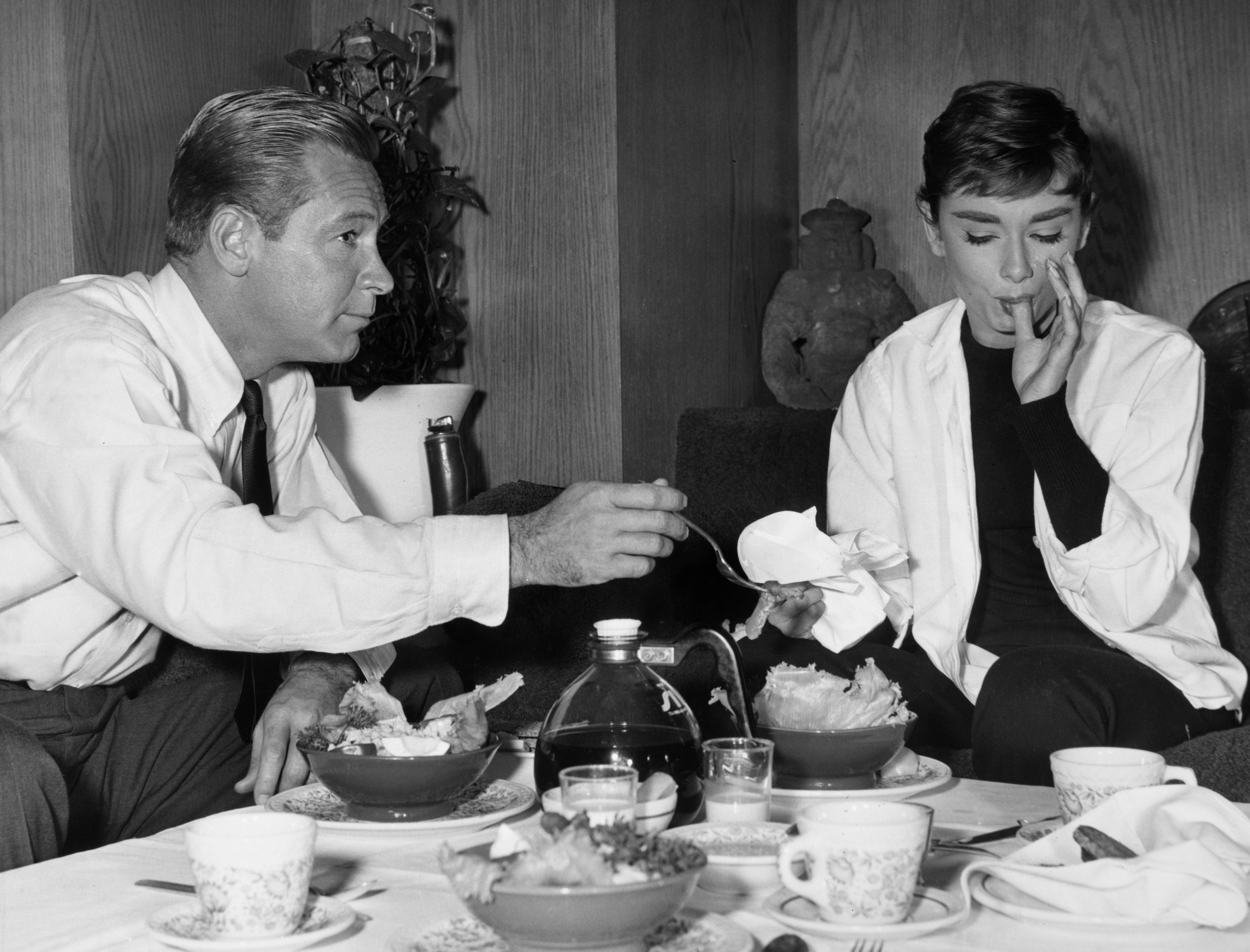 Two people having breakfast at a table. Audrey is elegantly dressed, seated with a thoughtful expression. Variety of food items on the table