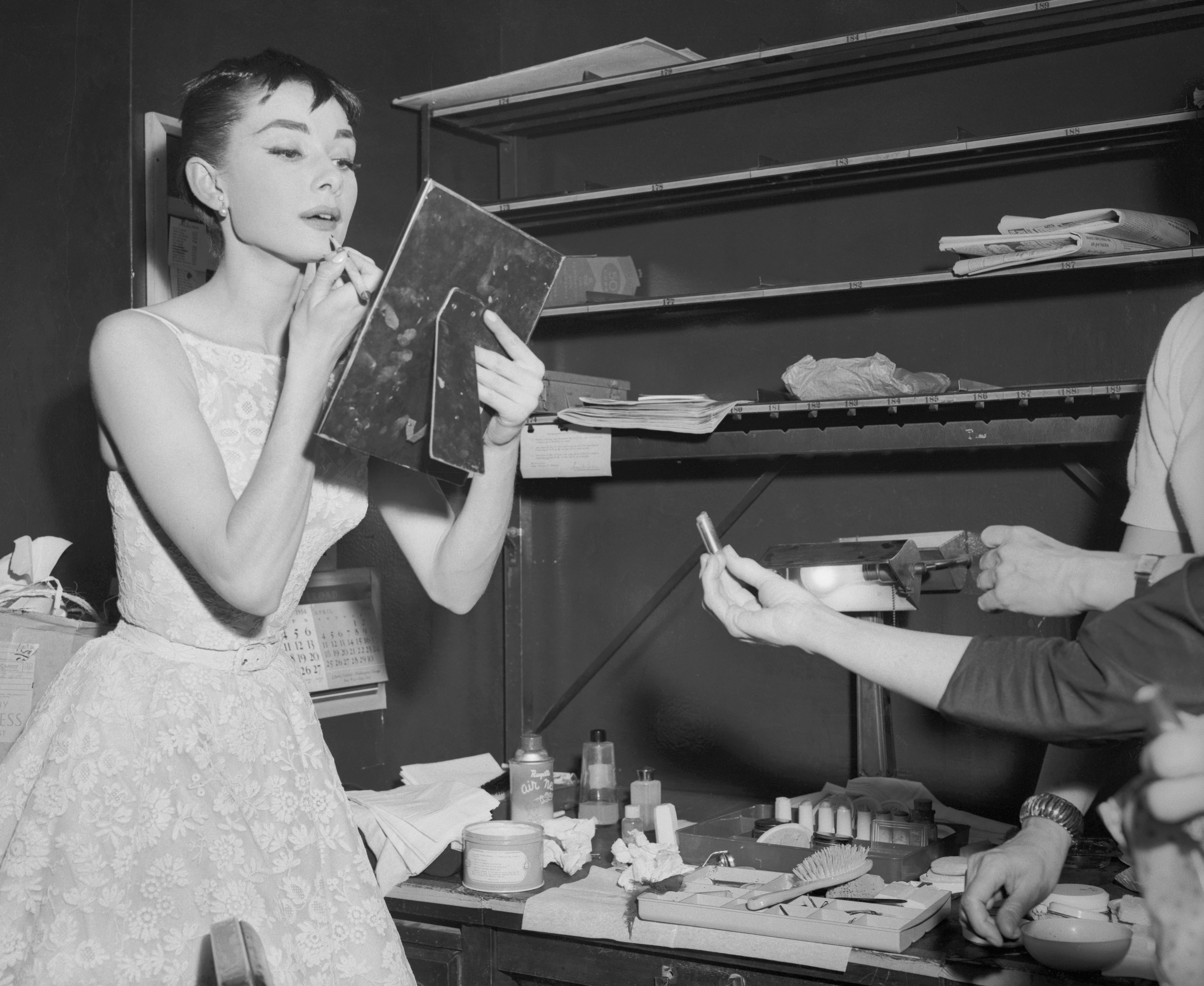 Audrey Hepburn in an elegant dress applies makeup using a handheld mirror backstage, surrounded by cosmetics and assisting hands