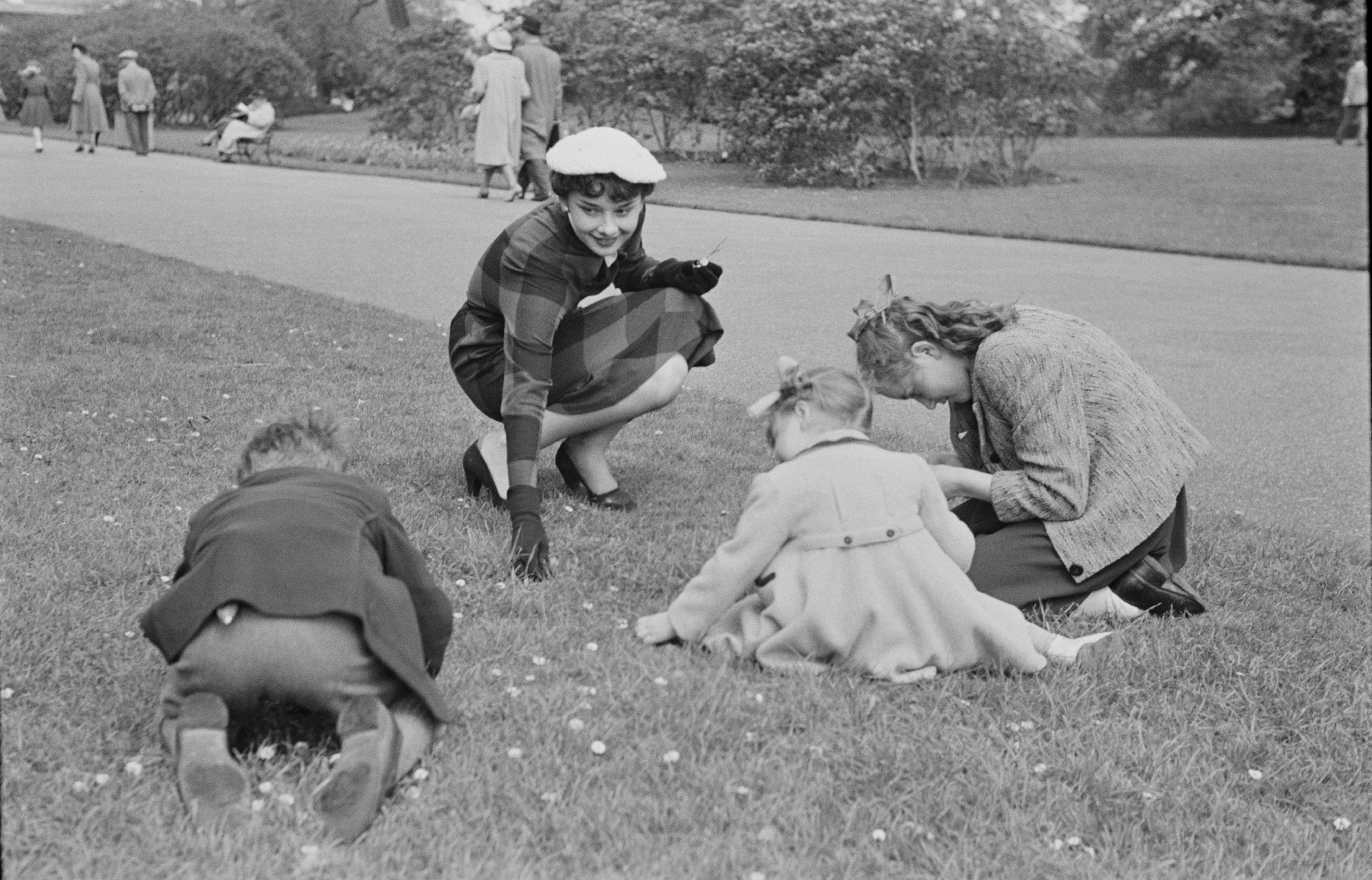Audrey Hepburn in stylish 1950s attire kneels on grass, playfully engaging with three children who are focused on the ground