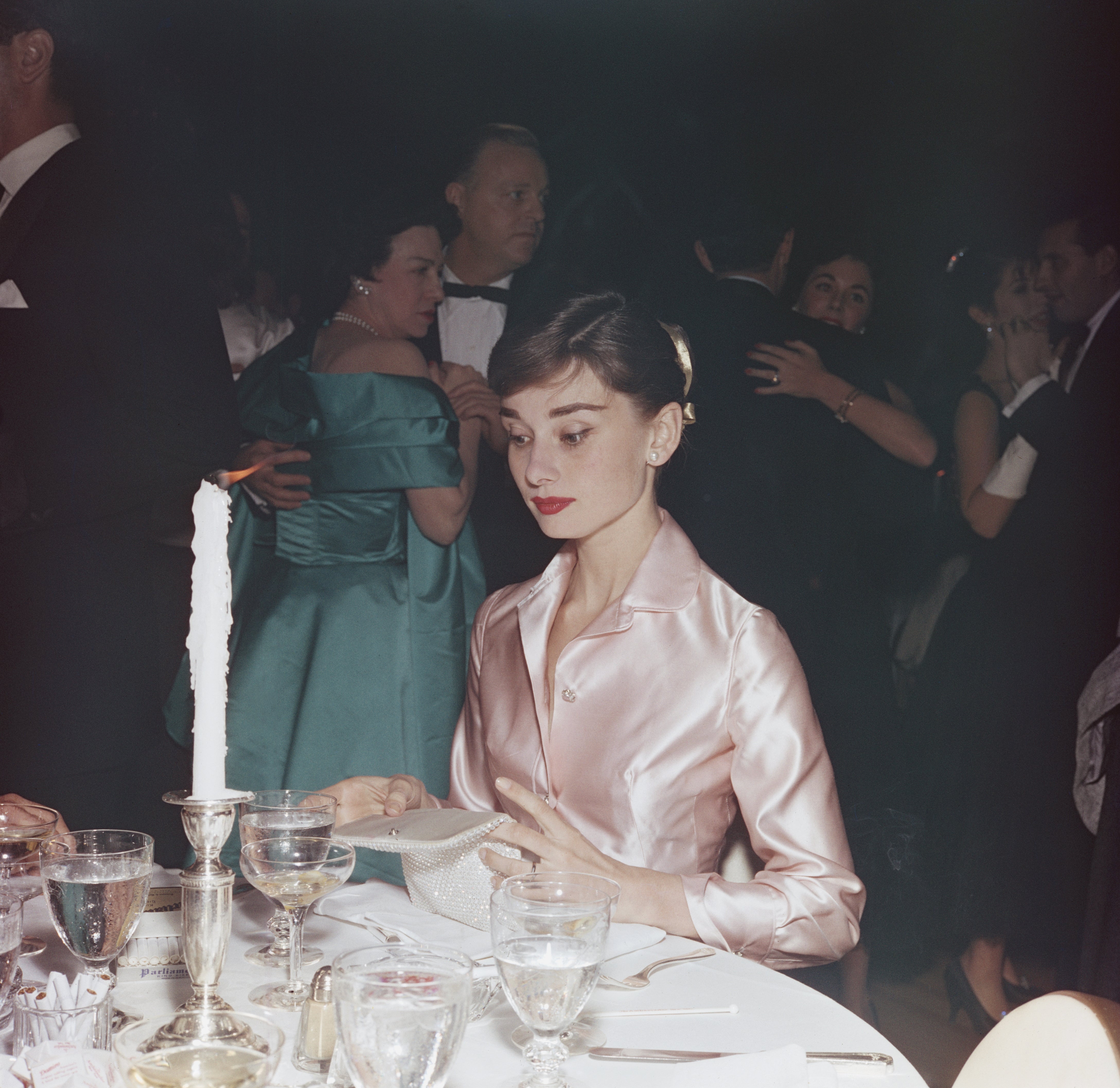 Audrey Hepburn in elegant satin gown seated at a formal event table, surrounded by other guests.