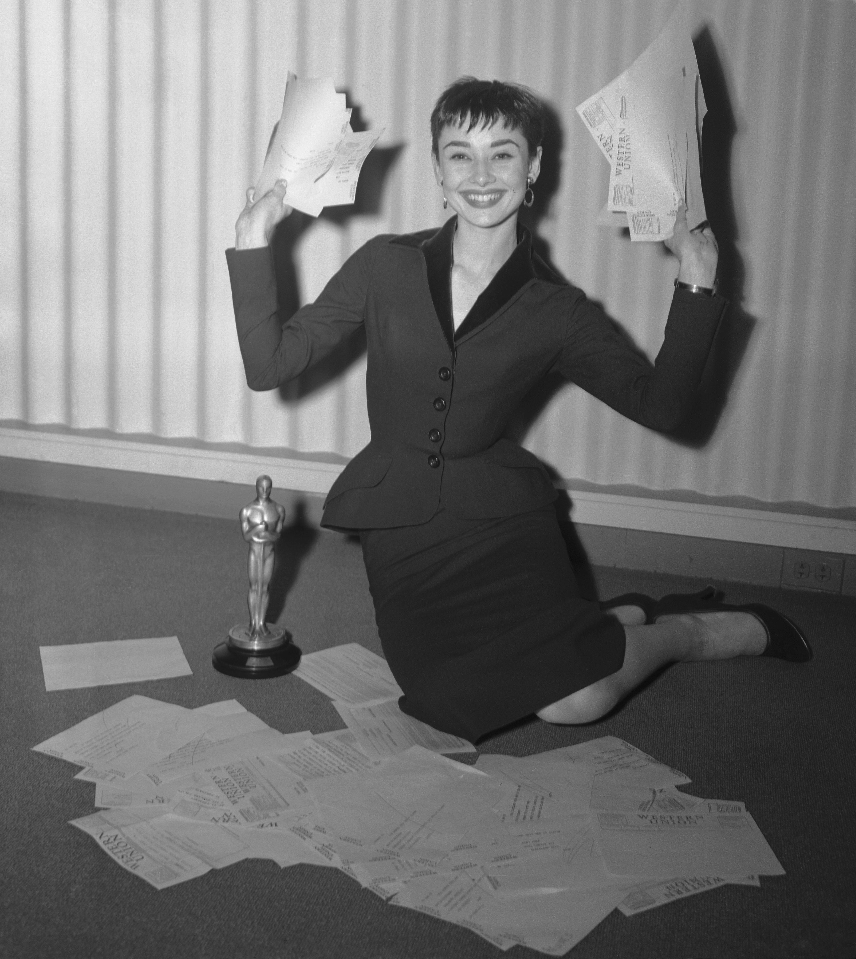 Audrey Hepburn kneeling, smiling, holding papers, surrounded by envelopes and a trophy resembling an Oscar statue
