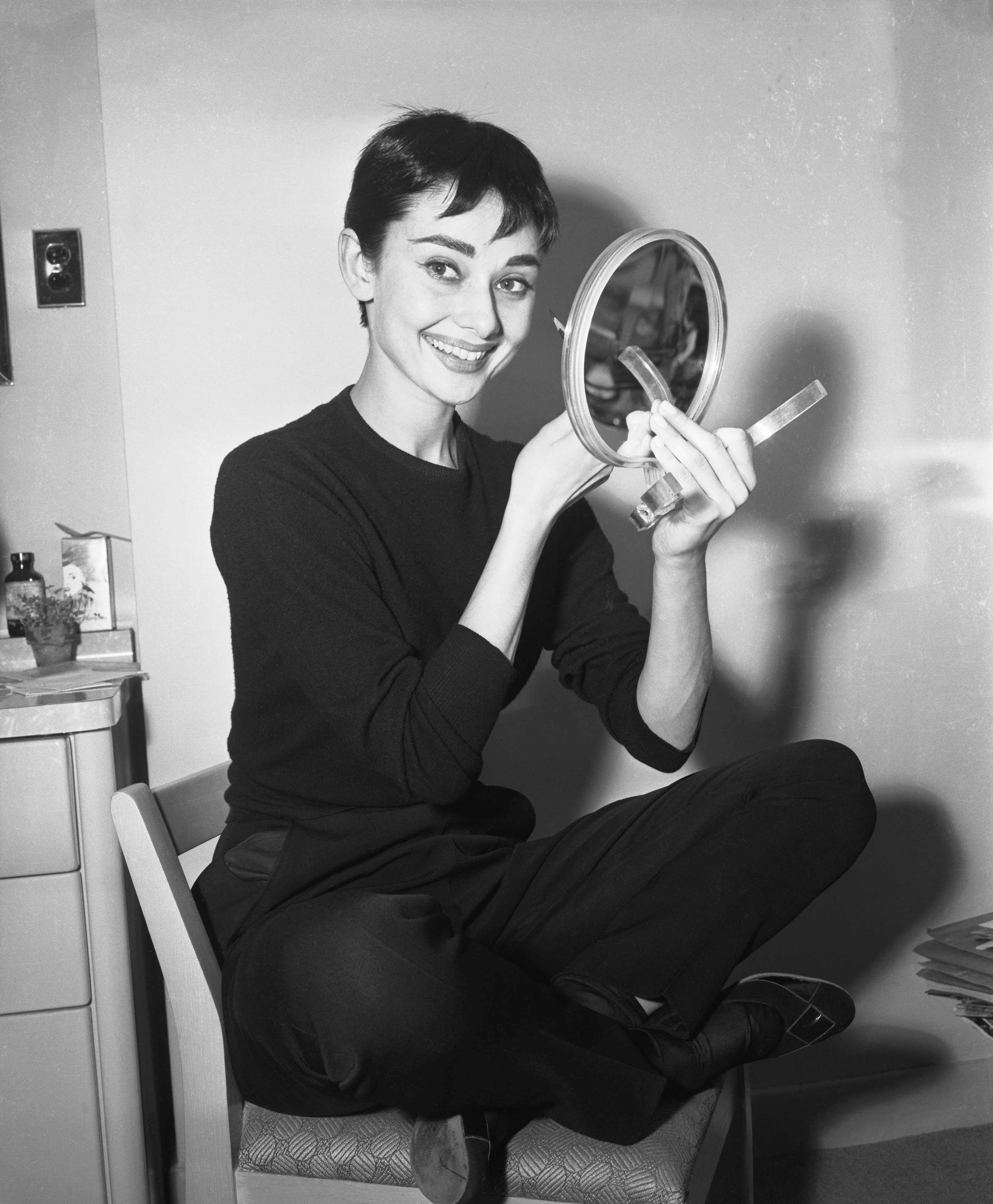 Audrey Hepburn seated, smiling, holding a handheld mirror while sitting cross-legged. They wear a simple, elegant style with a knit top and pants