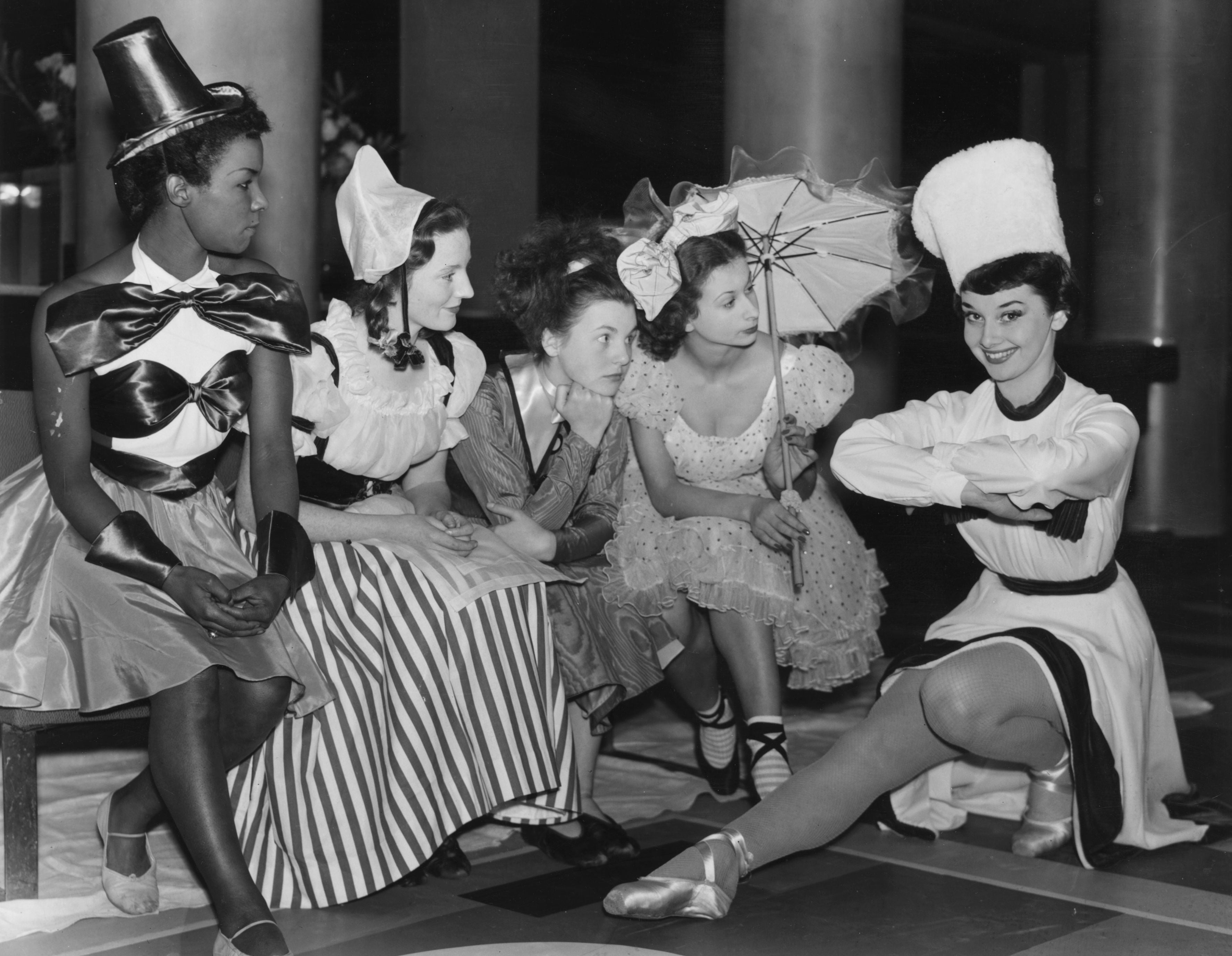 Performers dressed in elaborate, theatrical costumes with hats and accessories, seated and posing playfully, creating a lively and artistic scene