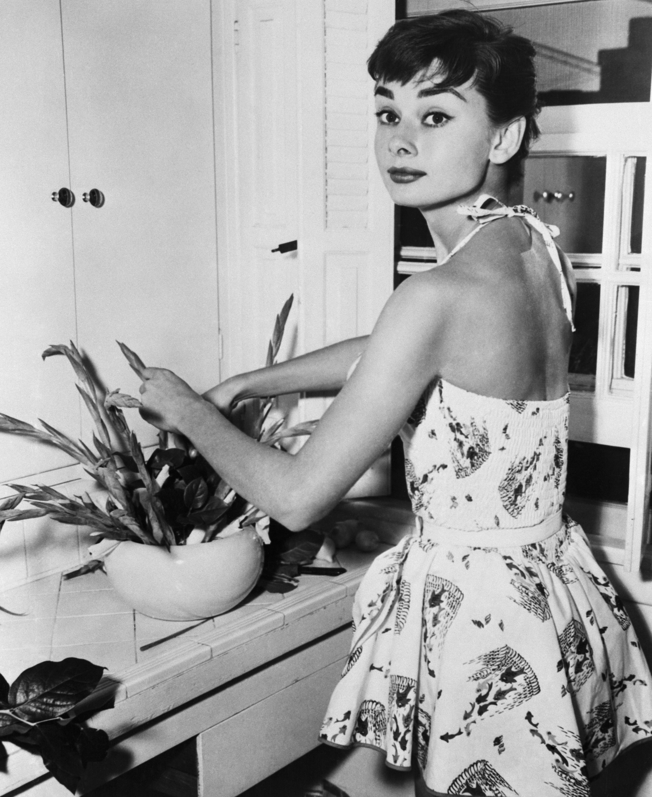 Audrey Hepburn in a sleeveless summer dress with a nature print, arranging plants in a bowl indoors