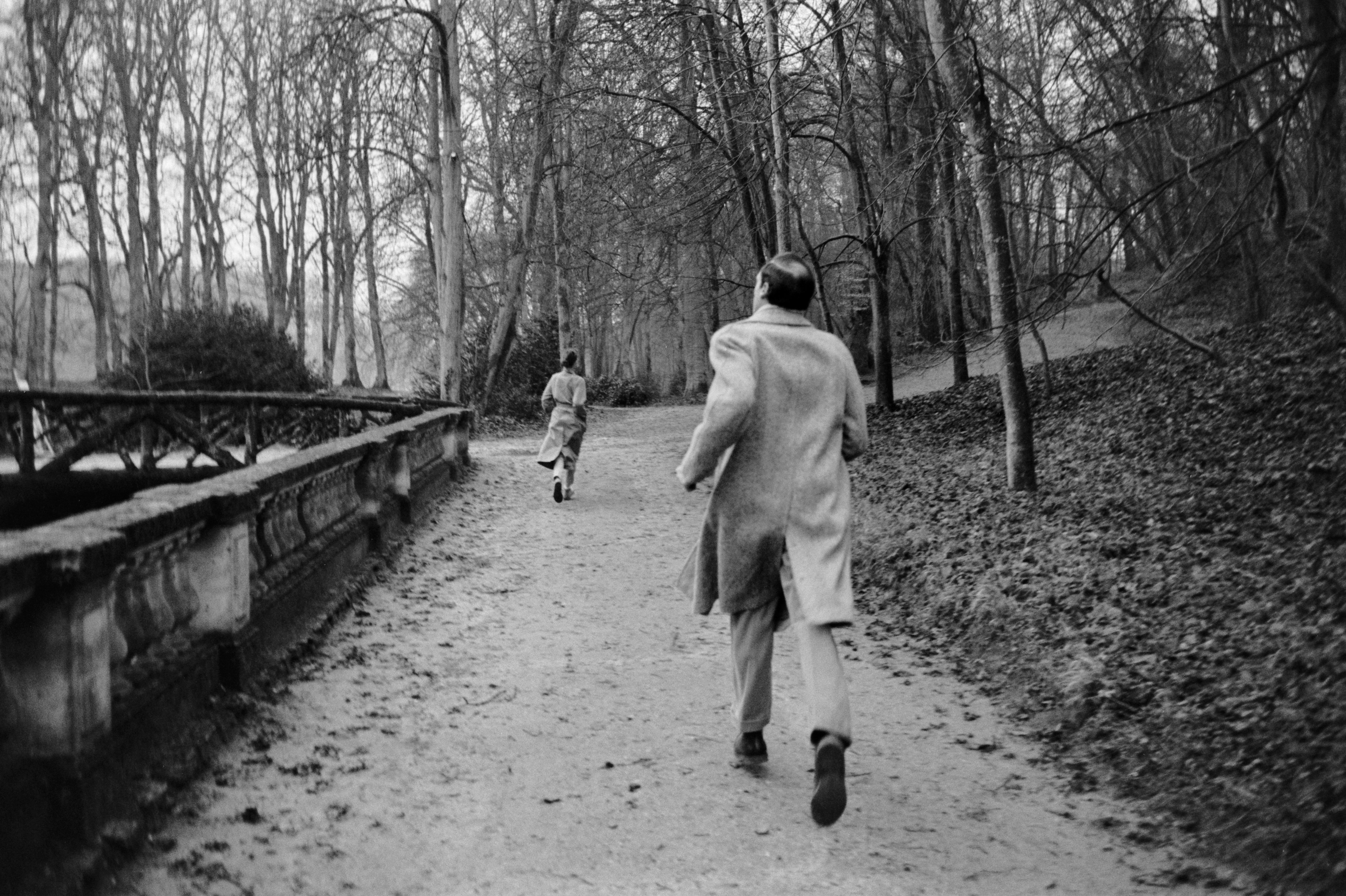 Audrey Hepburn runs along a tree-lined path in a park with Mel following closely behind, both dressed in coats
