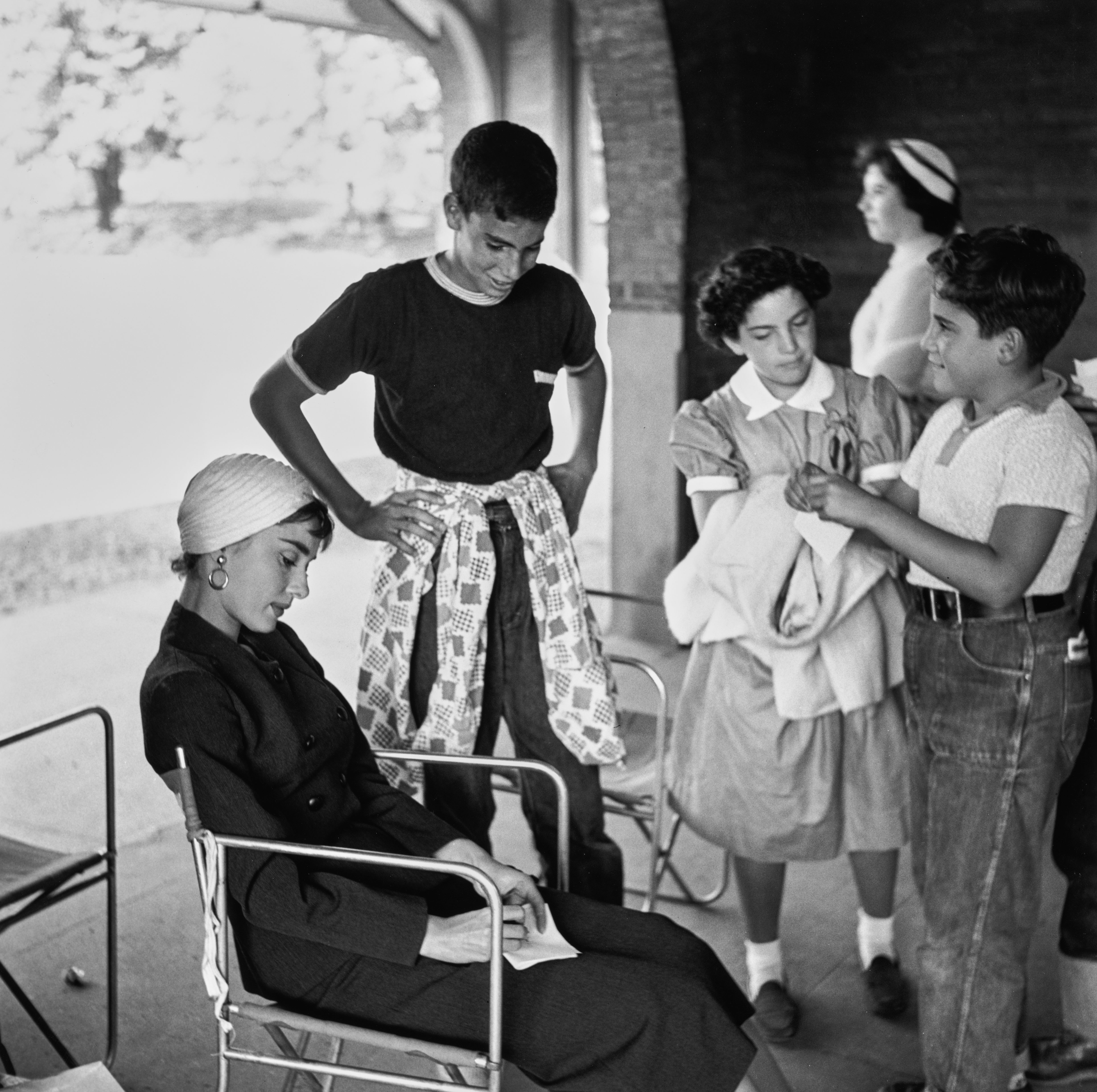 Audrey Hepburn sits on a chair, wearing a stylish headscarf and classic attire, while three children stand nearby, engaged in conversation