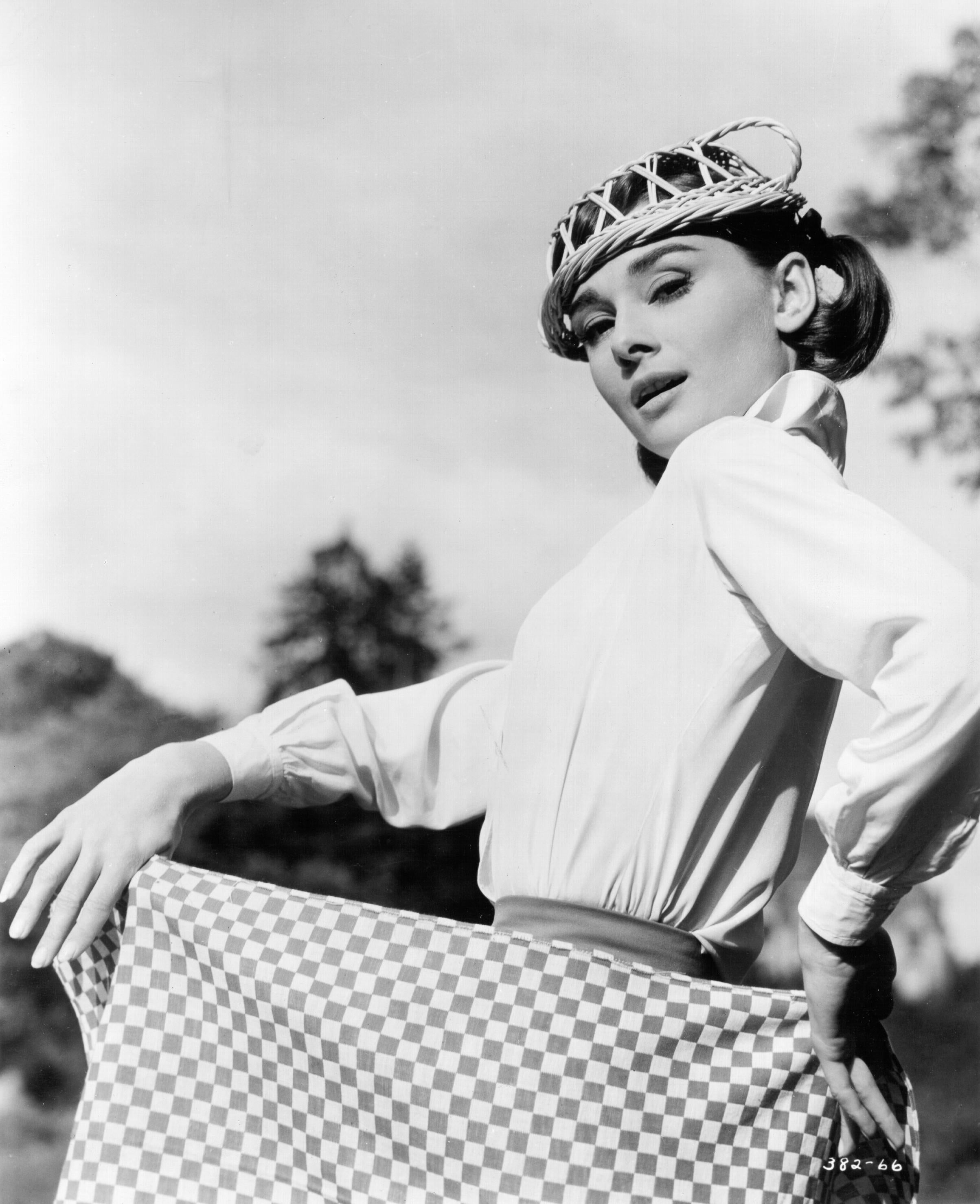 Audrey Hepburn in a classic photoshoot poses with a checkered dress and a stylish hat, evoking a timeless, elegant fashion sense