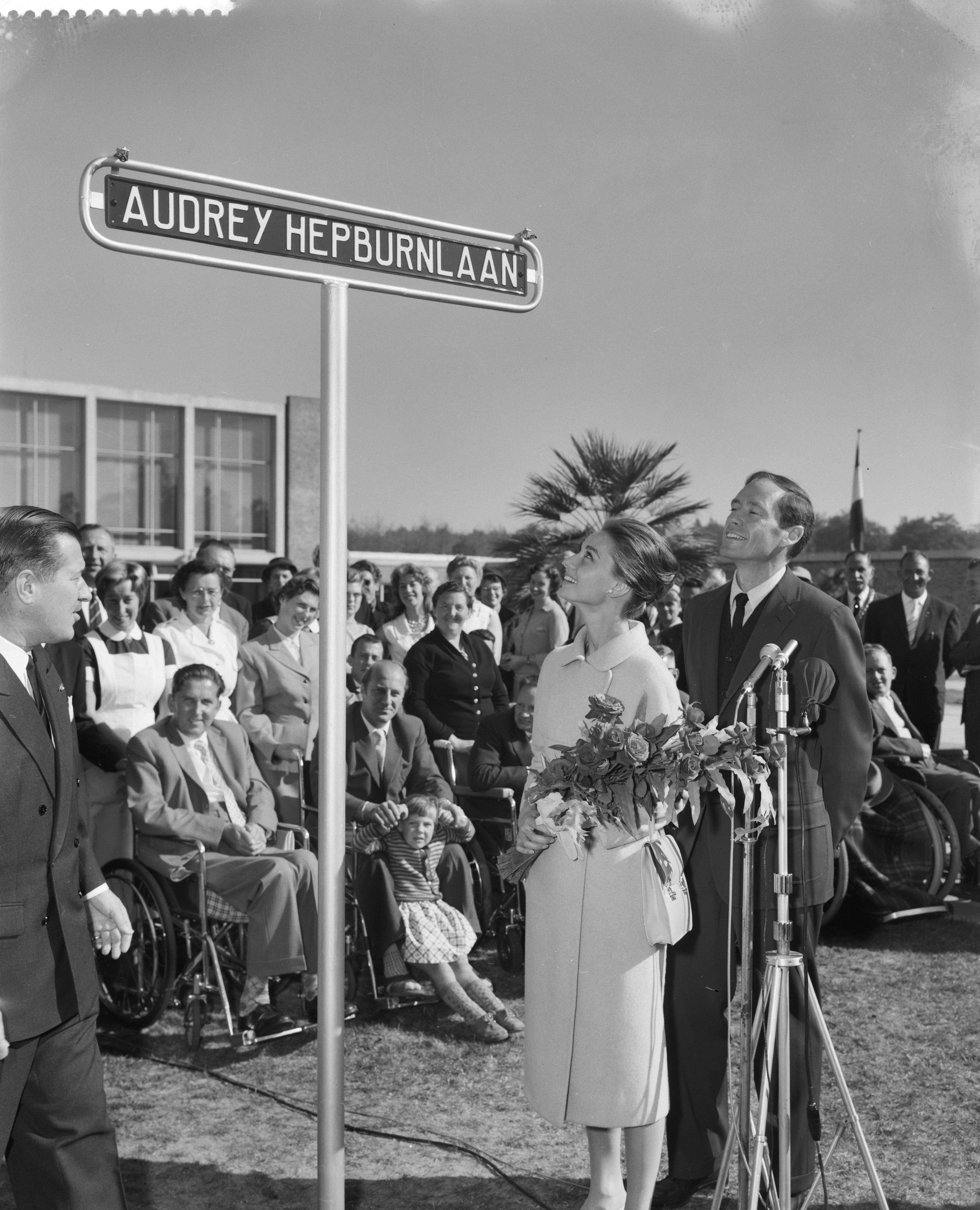 Audrey Hepburn stands in front of a street sign named after her, holding a bouquet, surrounded by a group of people, some in wheelchairs