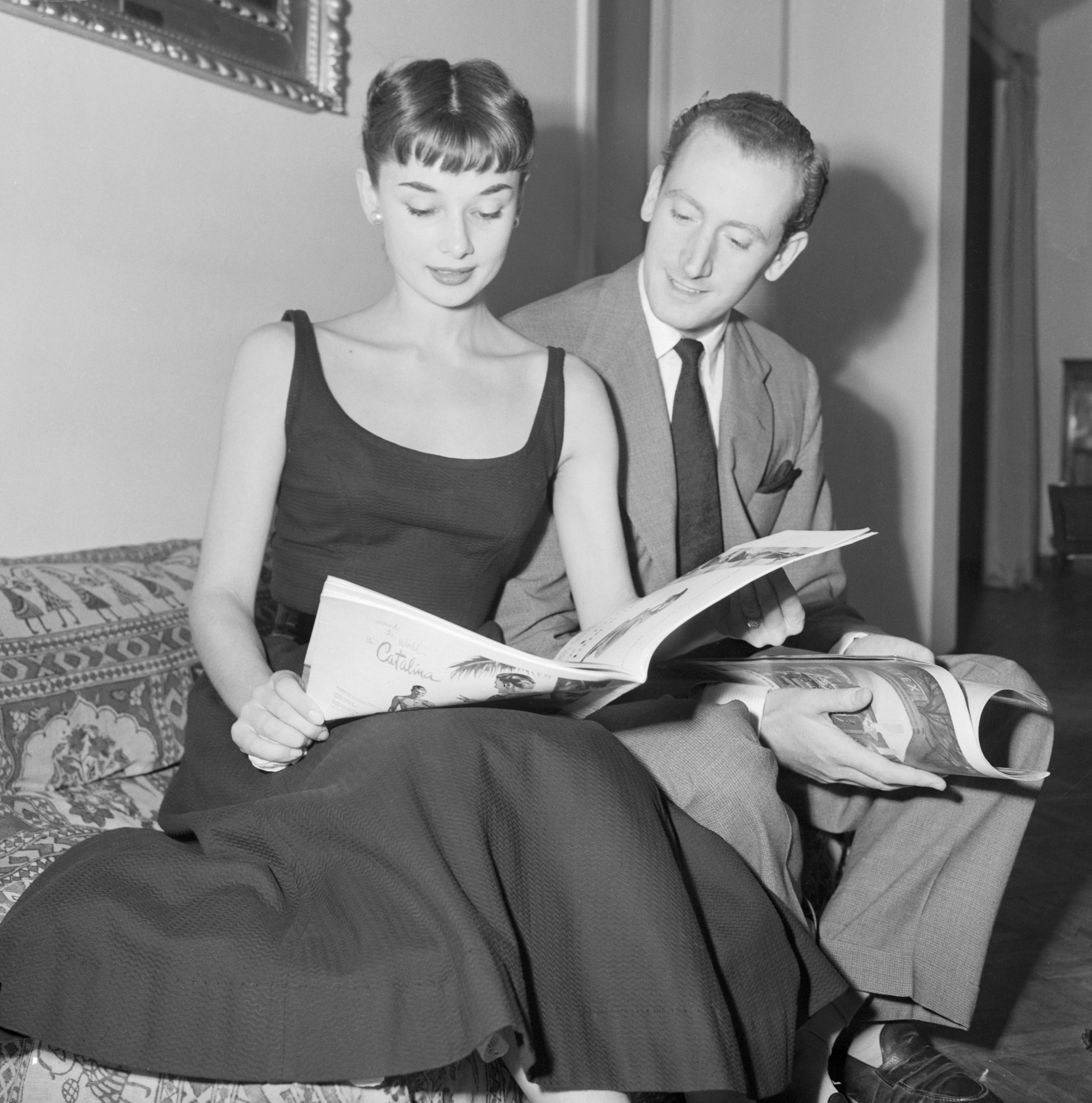Audrey Hepburn in a classic dress and James Hanson in a suit sit on a couch, looking at magazines together in a vintage setting
