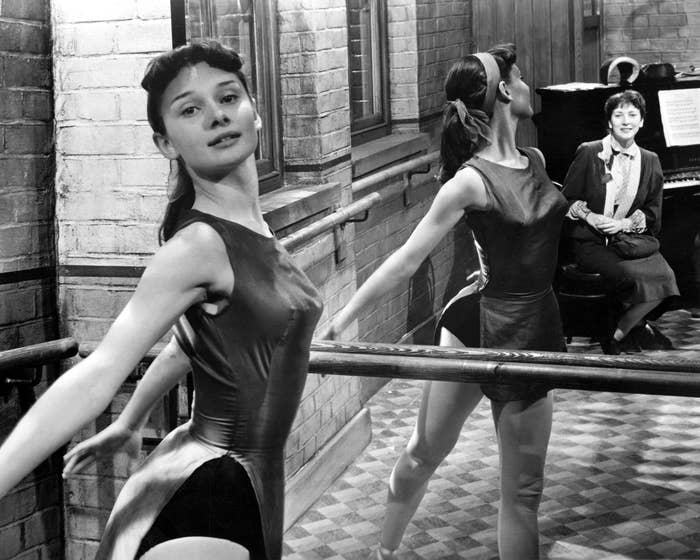 Audrey Hepburn practices at a barre in a studio, with an instructor nearby at the piano.