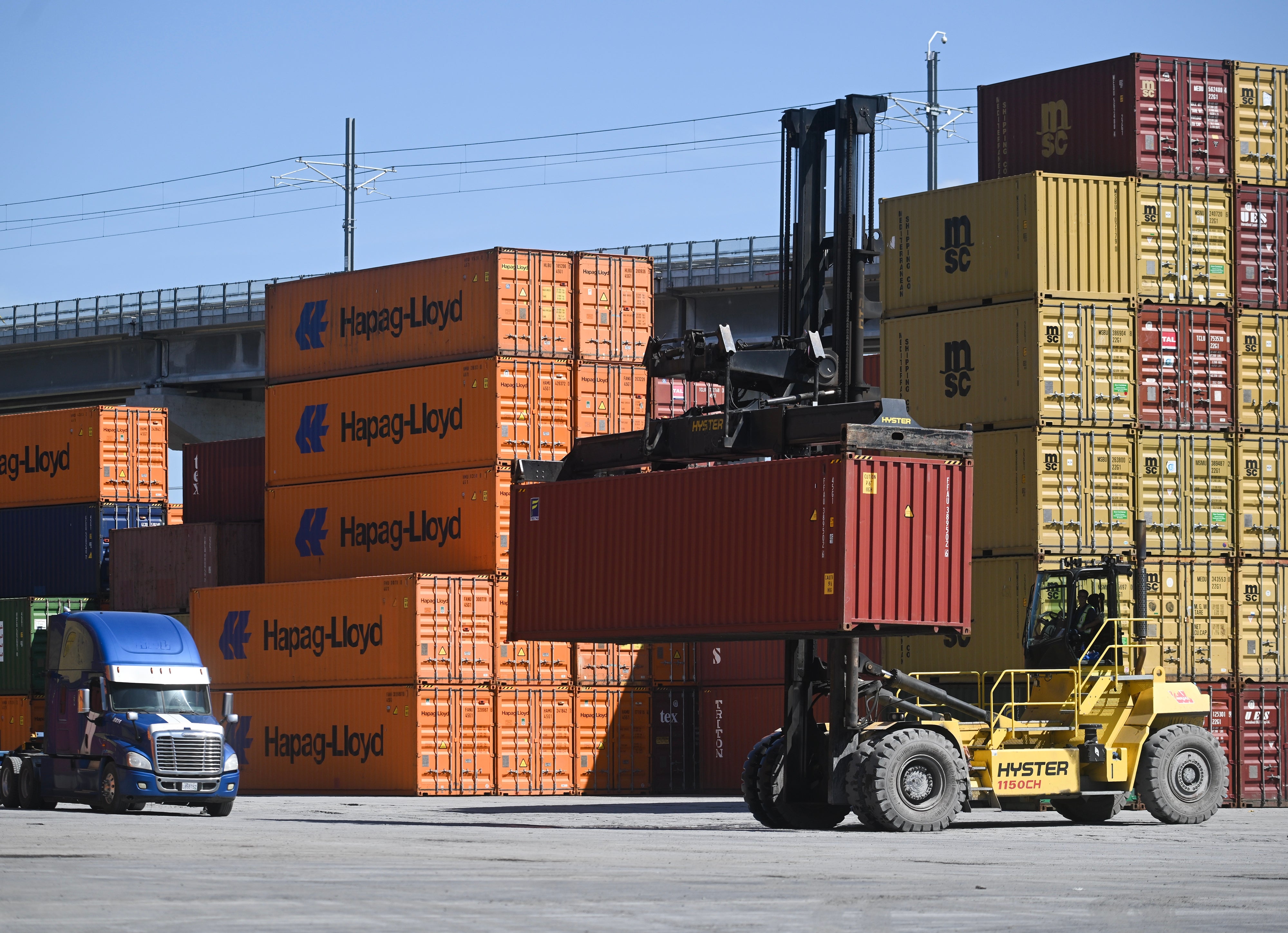 Gaffeltruck flytter en fraktcontainer i et travelt havneområde med stablede containere i bakgrunnen
