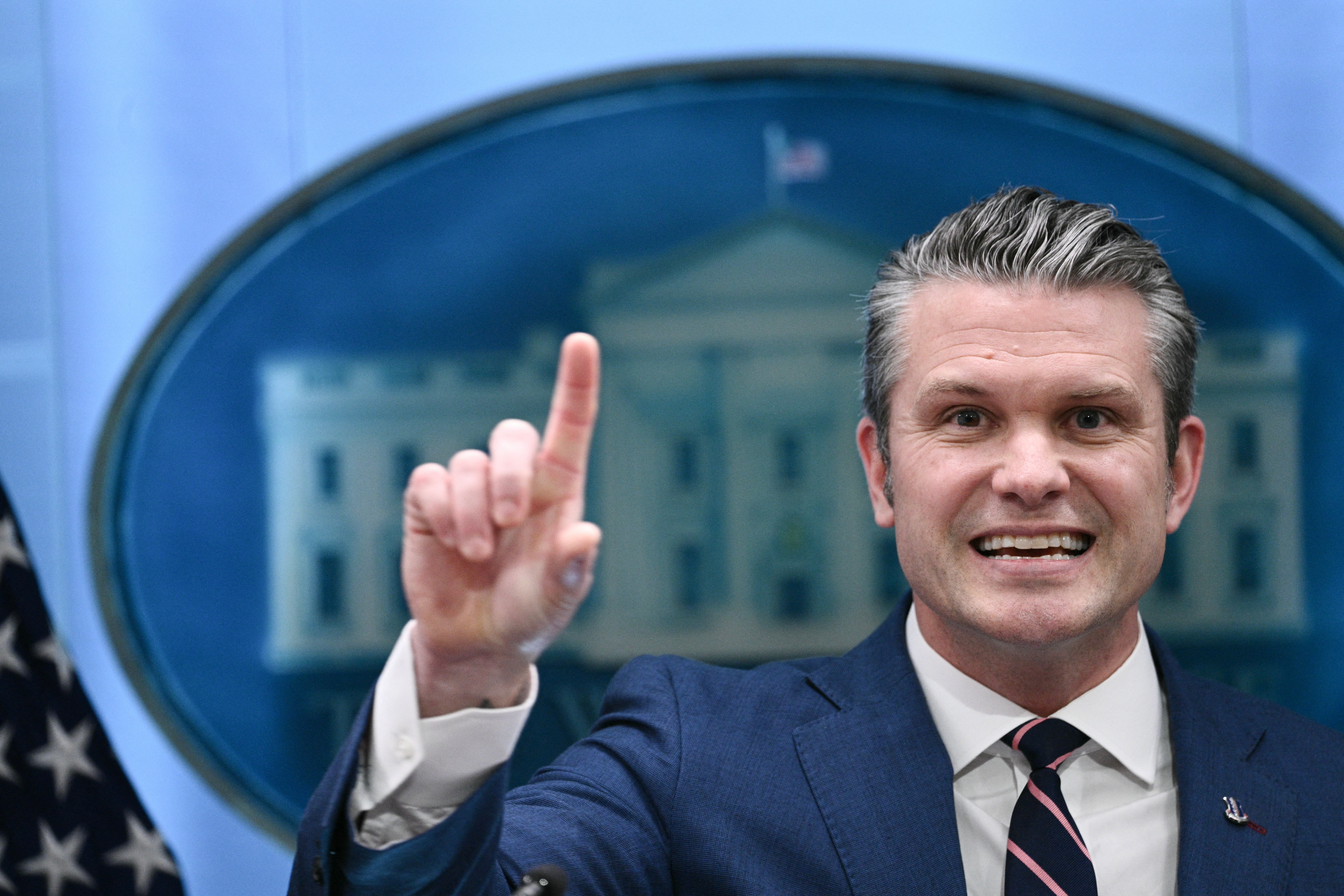 Smilende person i dress gester med én finger hevet foran en uskarp bakgrunn med en bygning