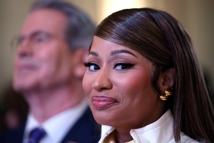 Person smiling at an event, wearing a white outfit with gold earrings, next to another person in the background