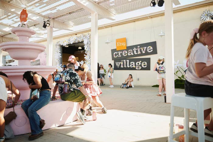 People sitting and walking around an outdoor event space labeled "Etsy Creative Village," showcasing various vendor booths and a pink fountain
