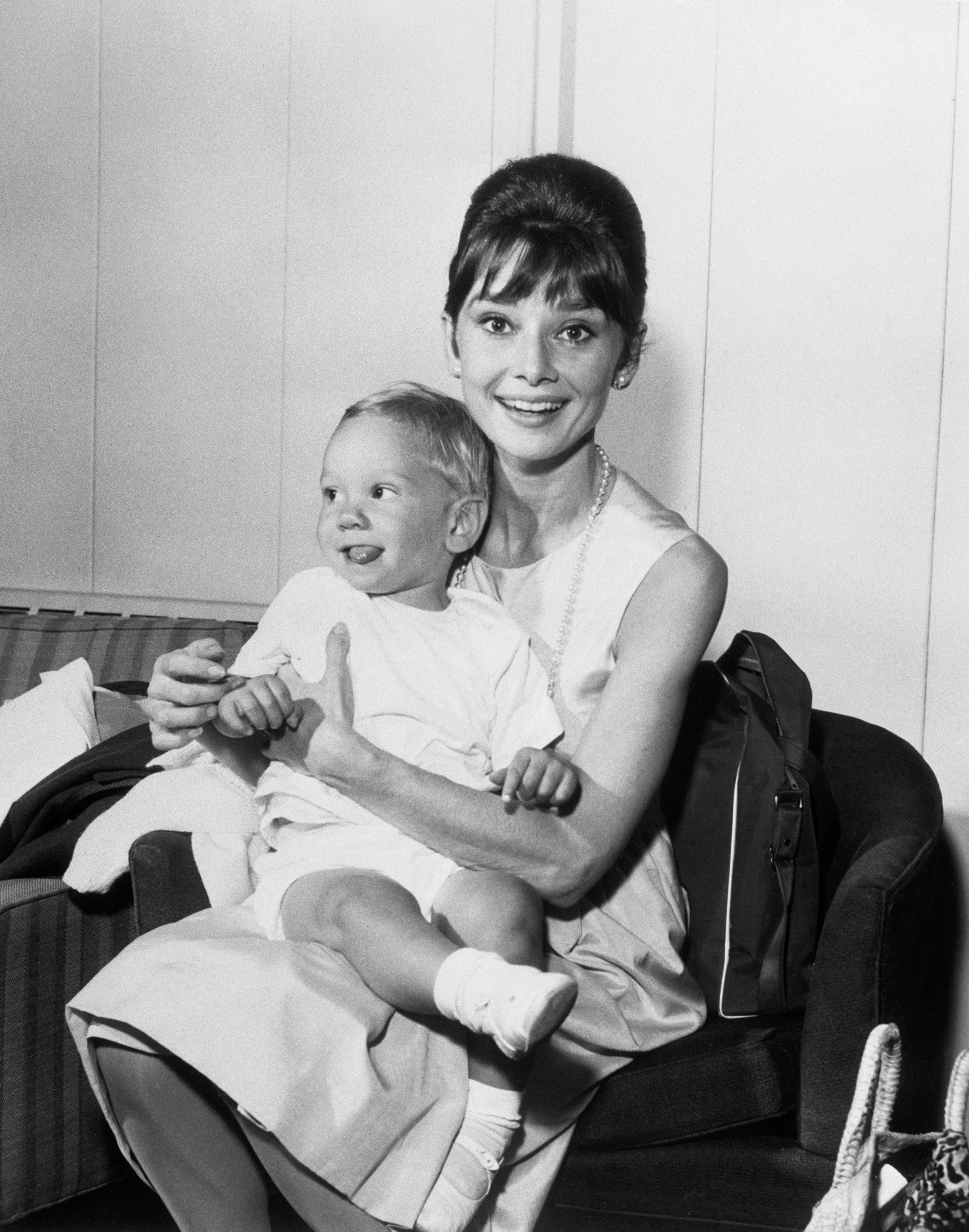 Audrey Hepburn with a classic updo and pearl necklace smiling while holding a young child seated on her lap. They are both seated on a sofa
