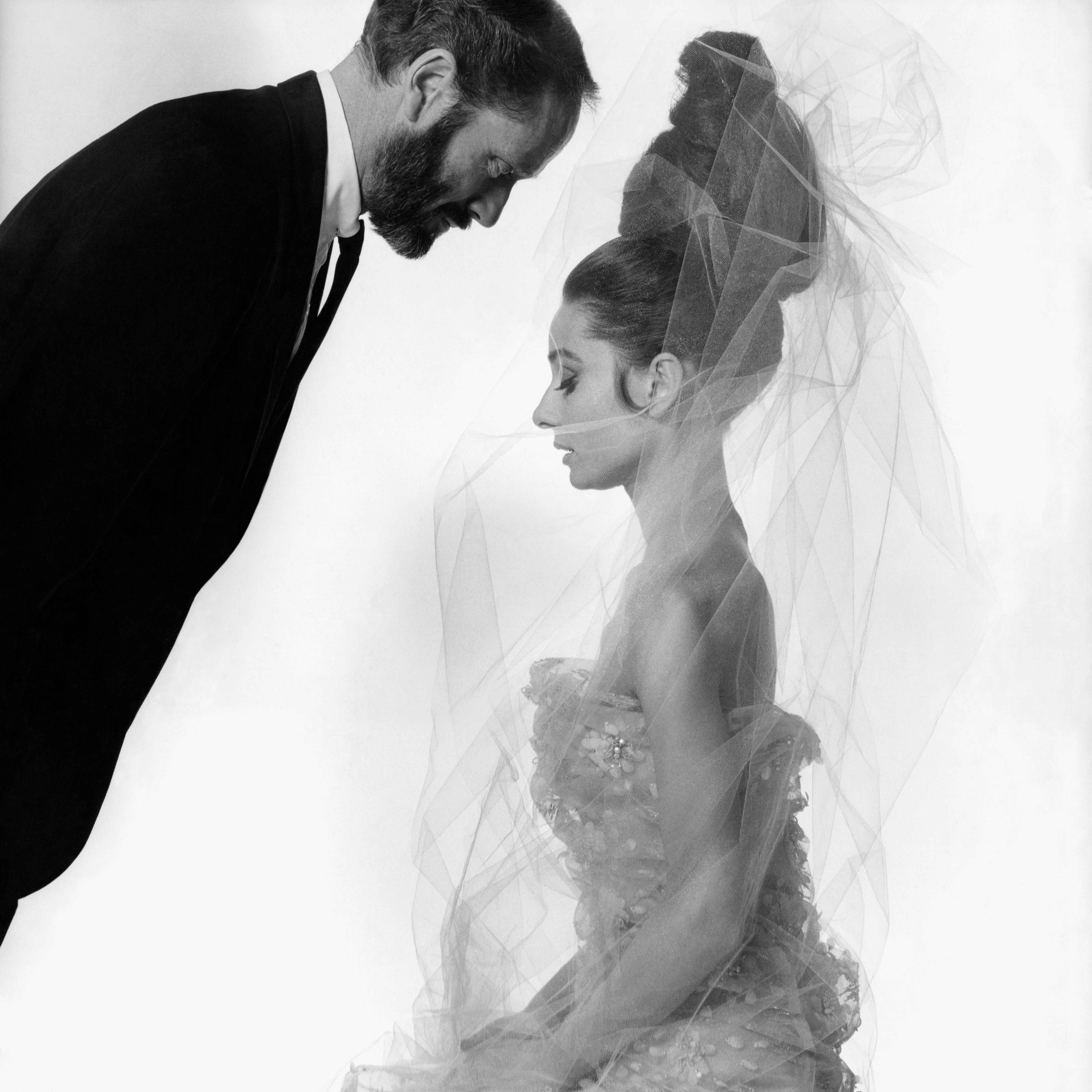 Audrey Hepburn in a textured dress and tall hairstyle sits, crowned with a veil, while another in a suit leans closely, creating a striking silhouette
