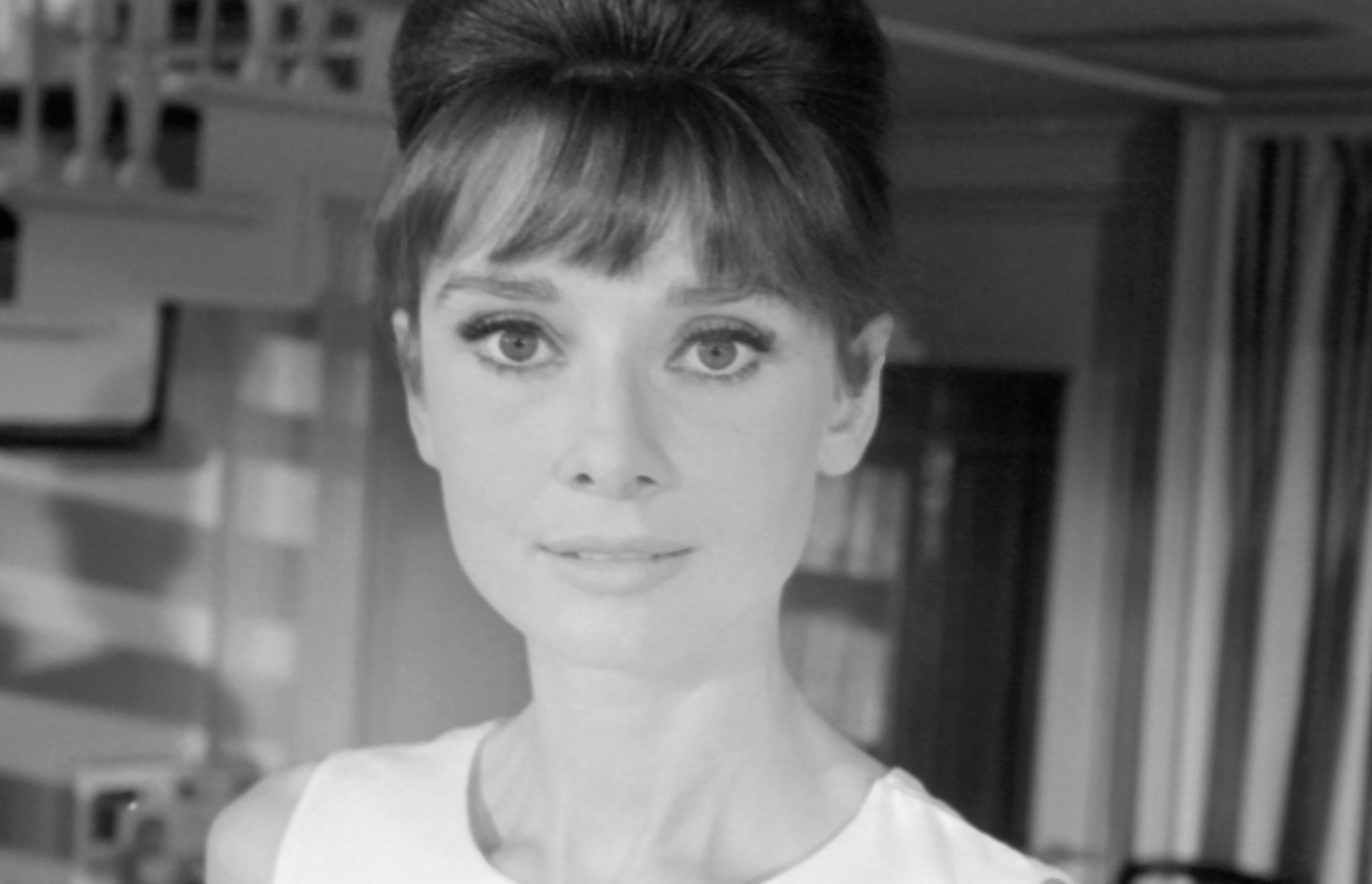 Audrey Hepburn with an elegant updo hairstyle and sleeveless top, looking directly ahead indoors