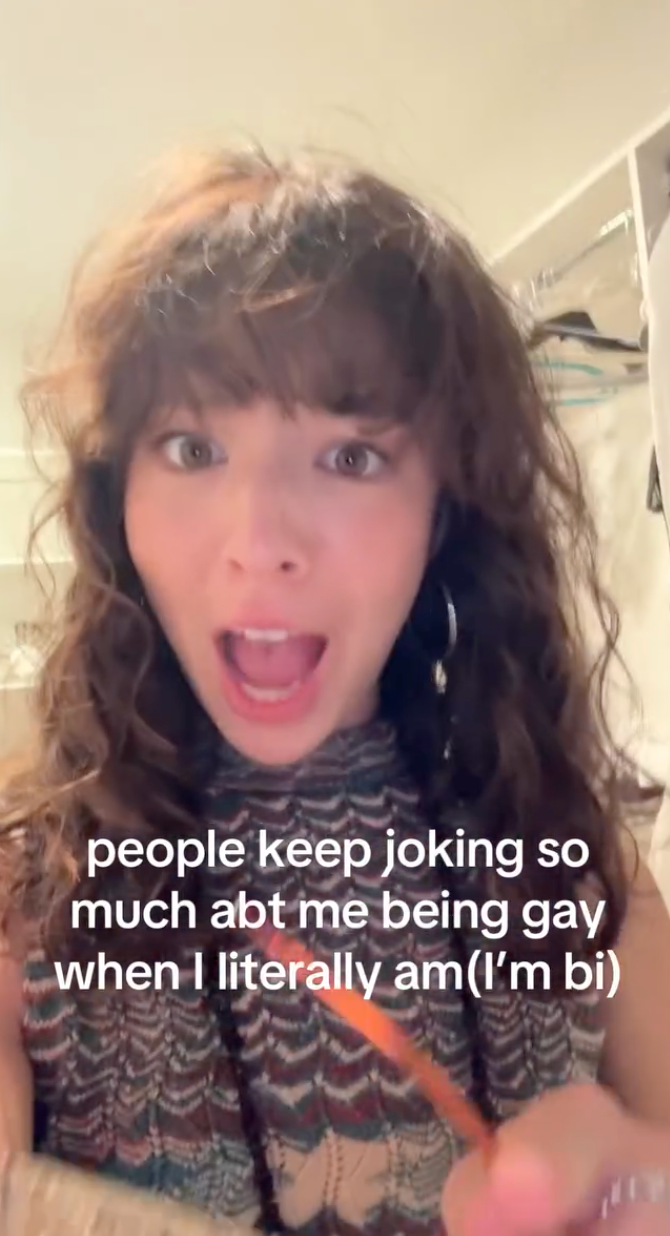 Person smiling with long curly hair, wearing a patterned top, with text saying people joke about them being gay when they are actually bi