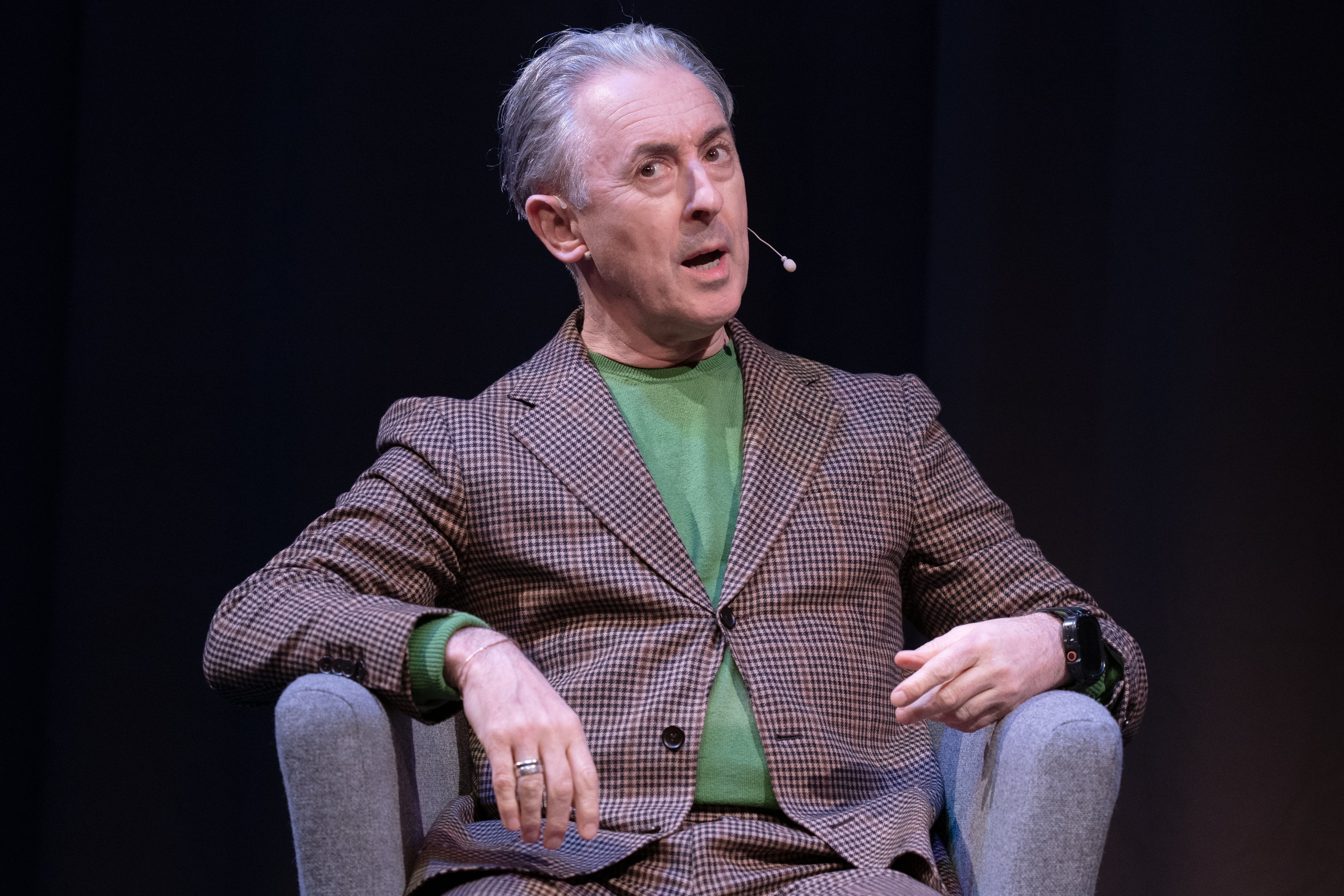 Person seated on stage, wearing a plaid suit and green shirt, speaking into a headset microphone