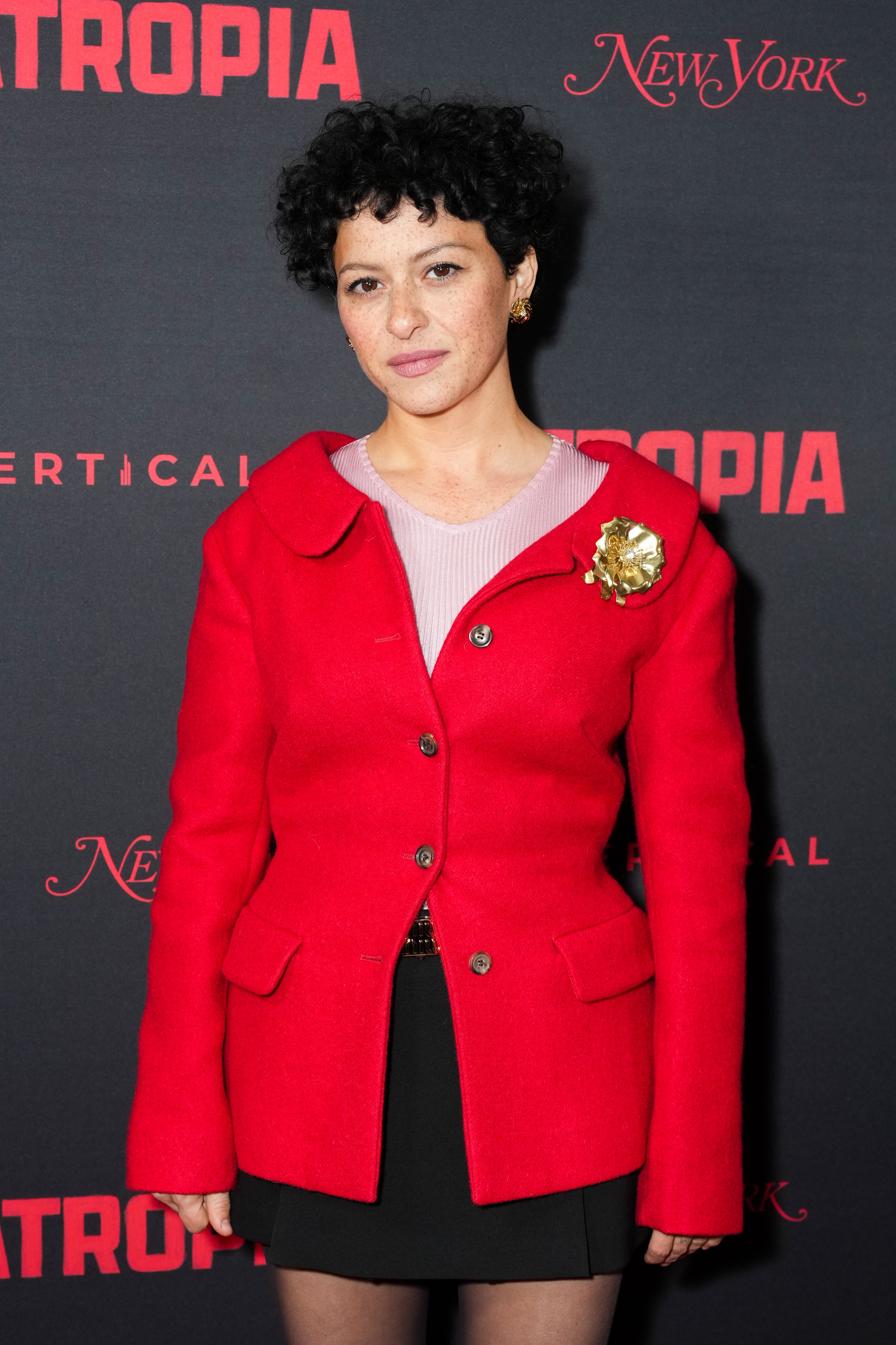 A person on the red carpet wears a stylish red jacket with a gold brooch, standing in front of a backdrop with promotional text