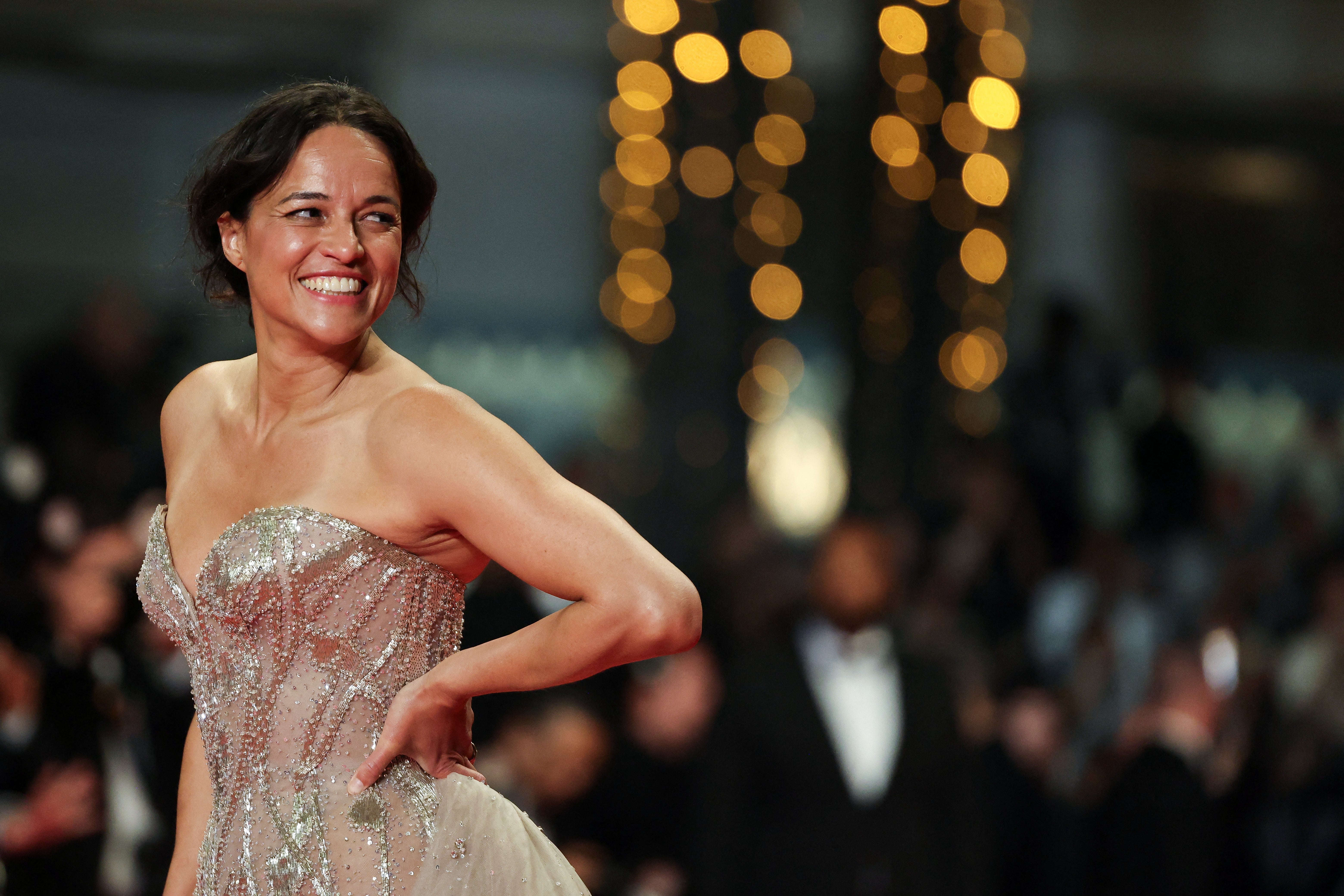 Person in elegant, strapless gown poses confidently on the red carpet, smiling brightly in a lively atmosphere with blurred lights in the background