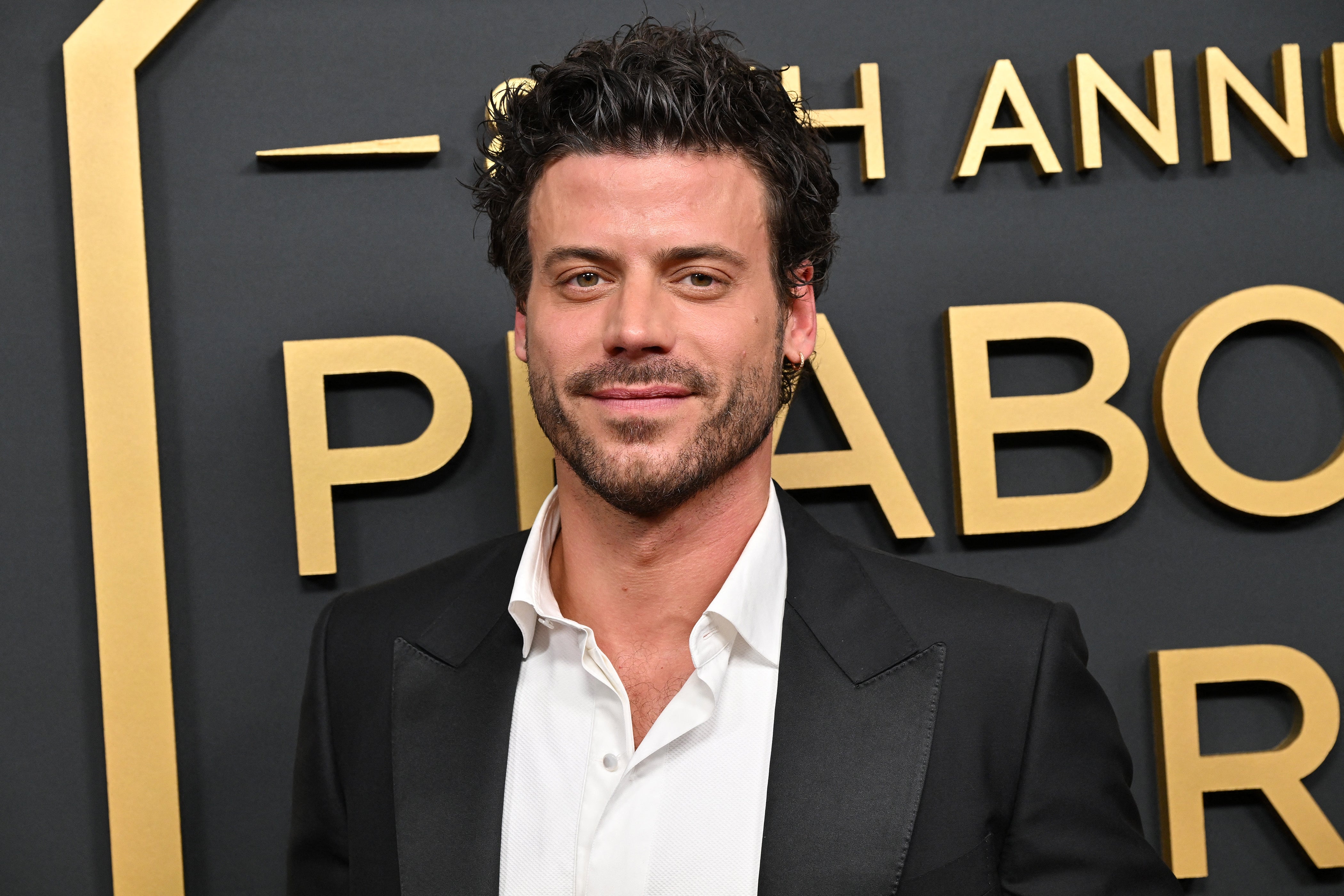 A person stands on the red carpet, wearing a classic black suit over a white shirt, in front of event signage