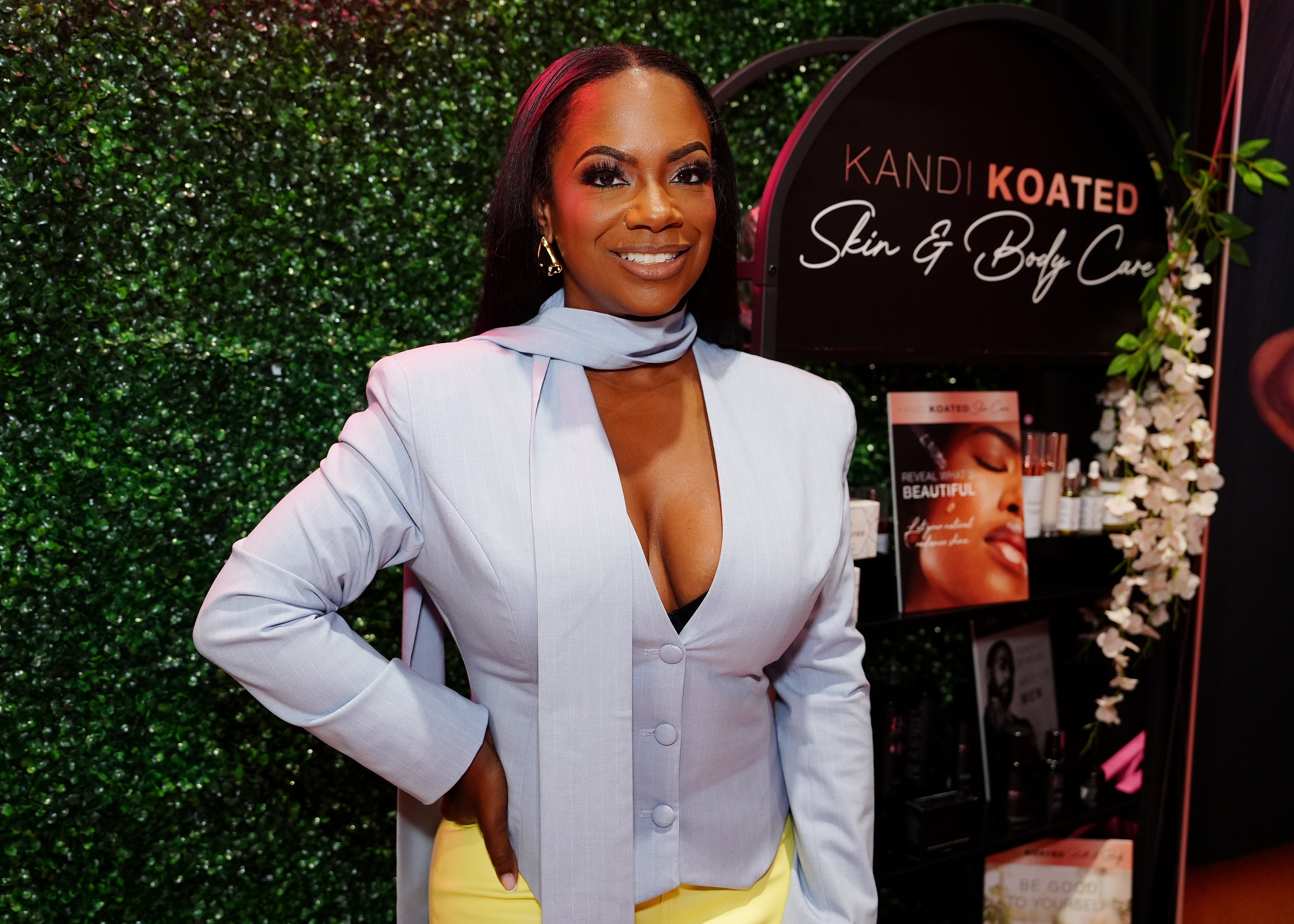 Person in stylish suit with scarf stands confidently at a skincare event for "Kandi Koated Skin &amp;amp; Body Care."