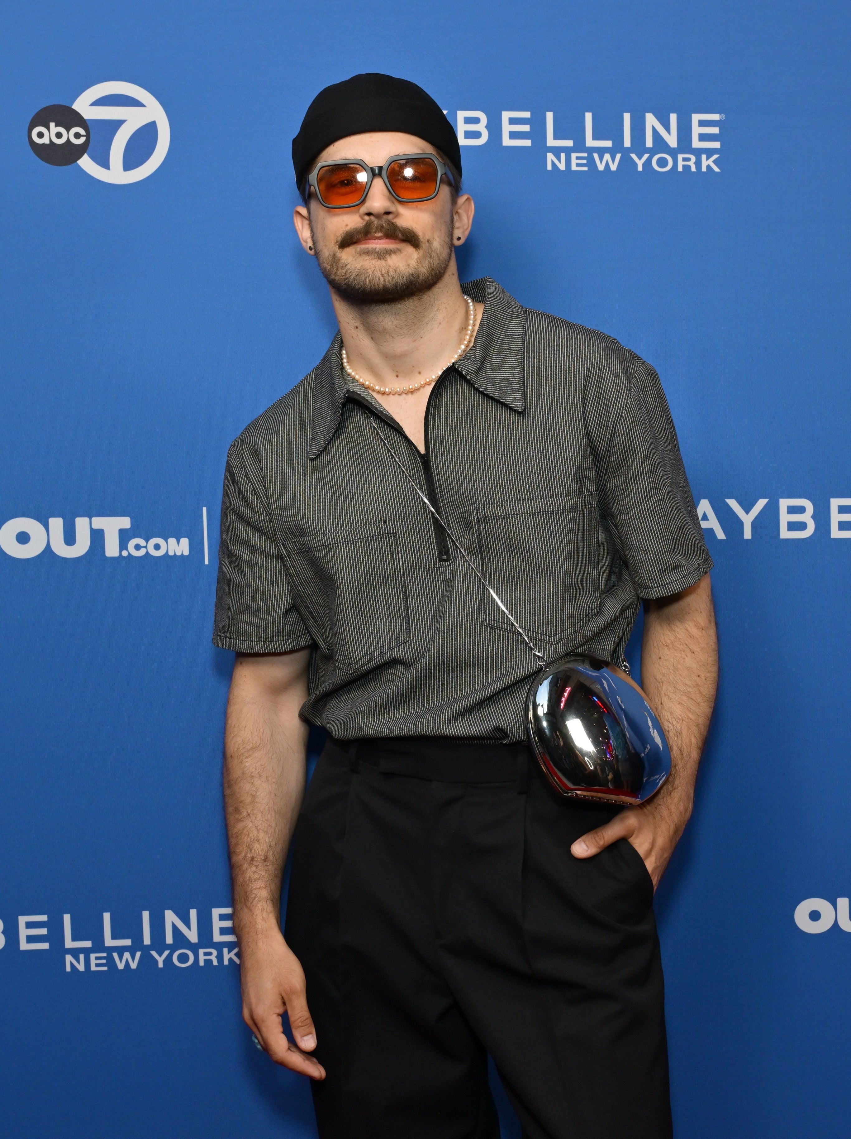 Person on press line wearing a cap, sunglasses, short-sleeve shirt, and high-waisted pants with a unique bag. Background has logos and text