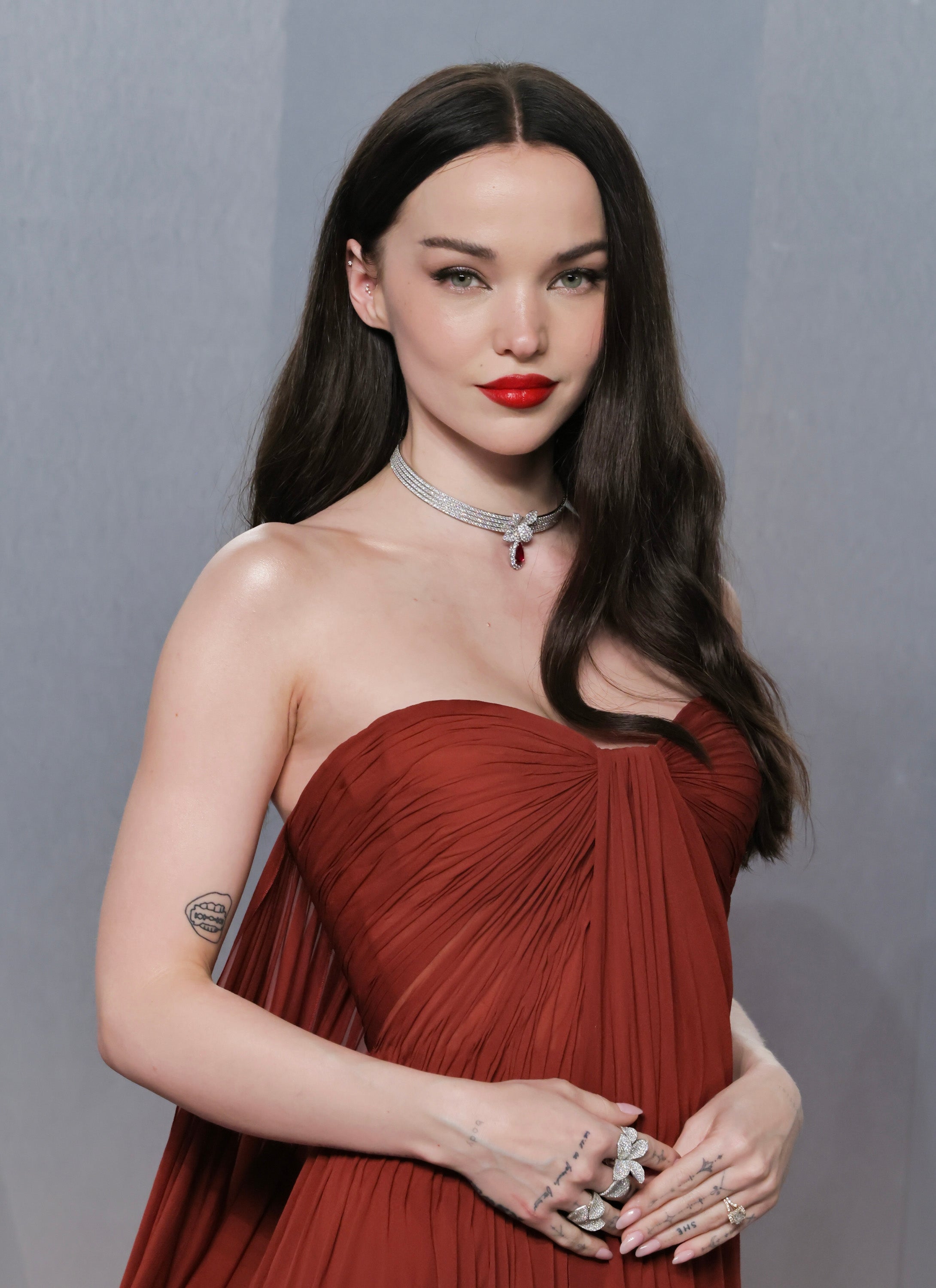 Person in elegant strapless gown with long hair and red lips, adorned with a choker necklace and multiple rings, stands poised for a photo