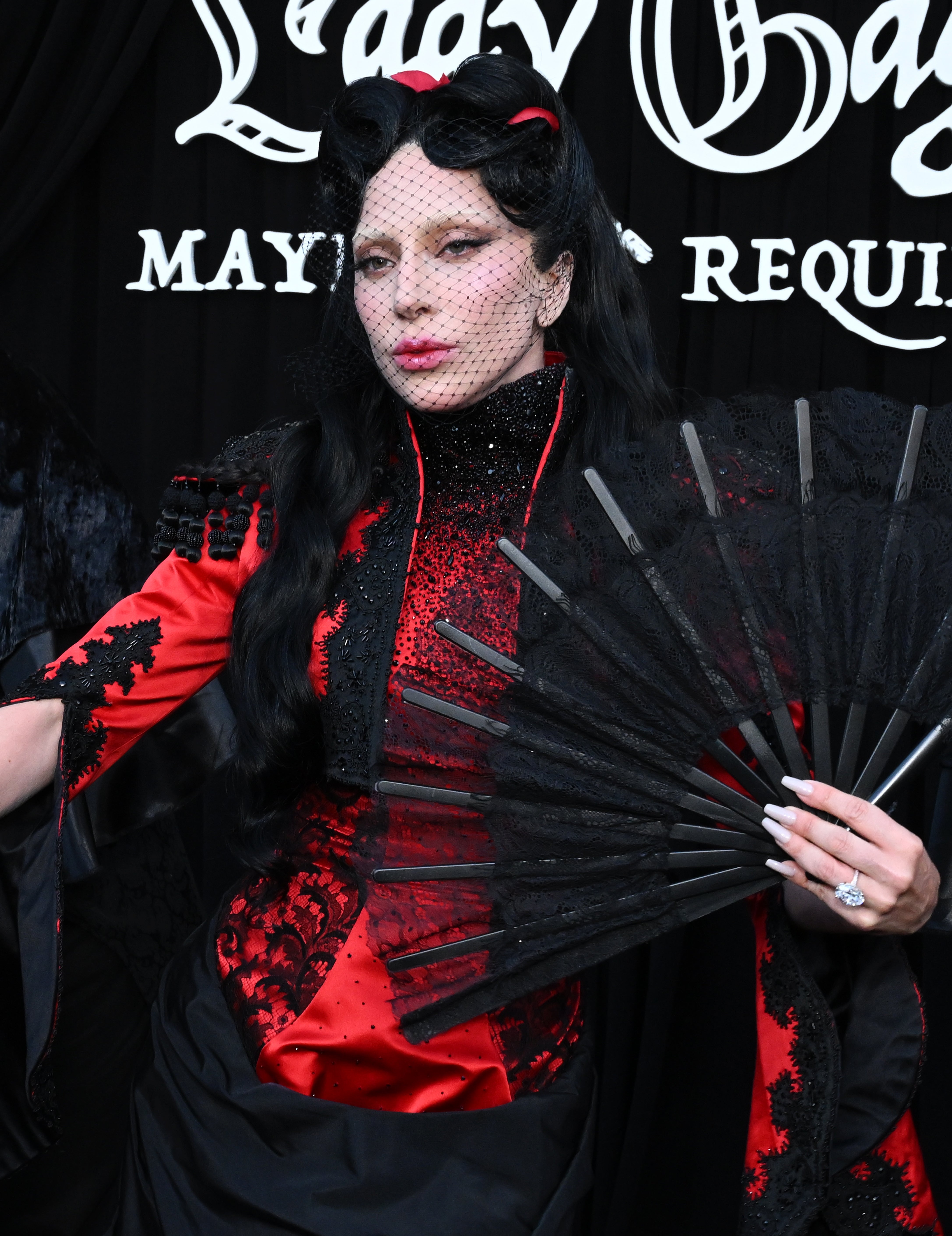 Person wearing an avant-garde outfit with lace details, holding a large black fan, under a sign with cursive writing