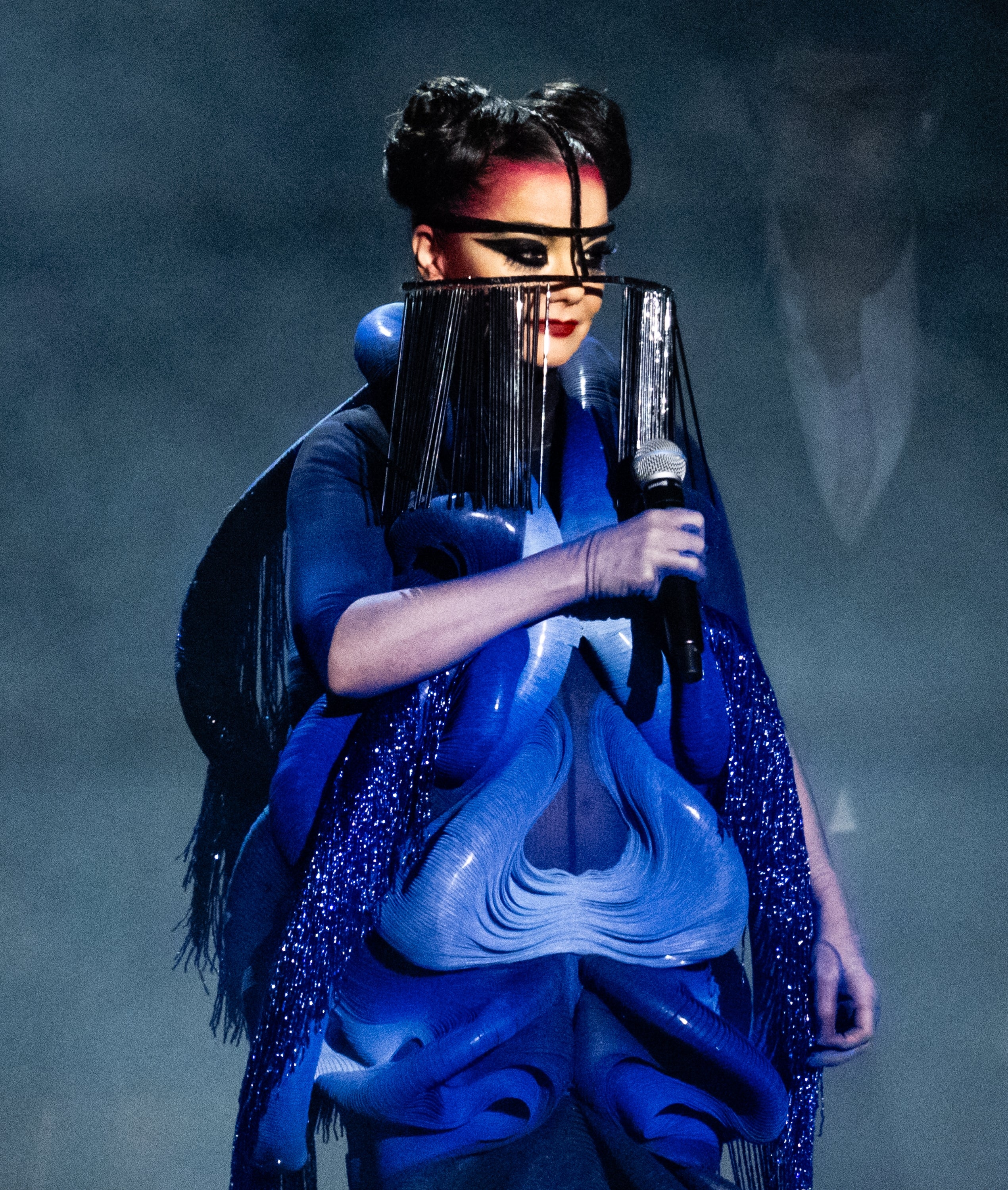 Person wearing avant-garde, futuristic outfit with elaborate headpiece, holding a microphone, on stage