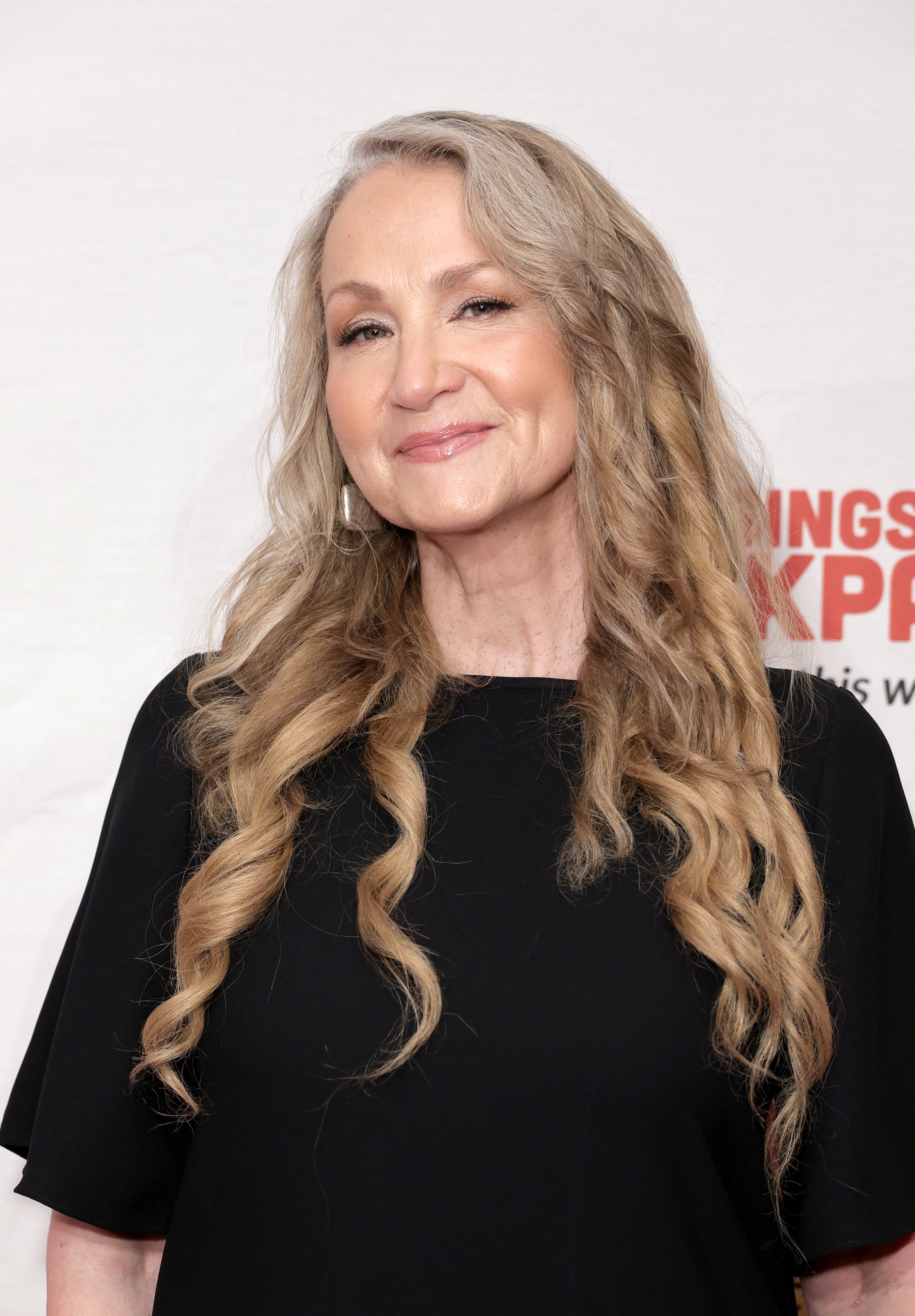 Person with long wavy hair in a simple, elegant black outfit, smiling at a public event