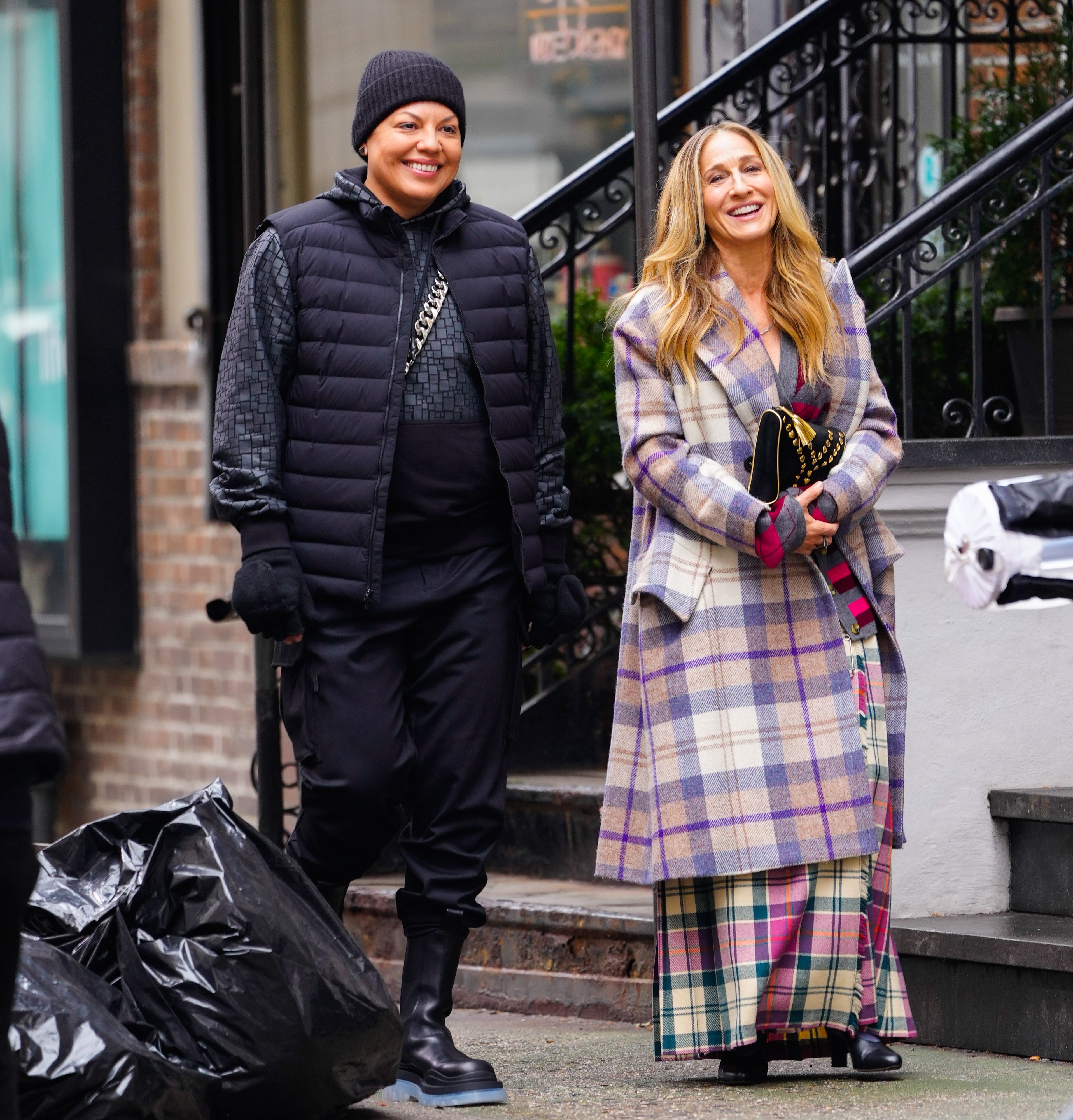 Two people smiling outdoors. One wears a beanie and puffer jacket; the other wears a stylish plaid coat over a vibrant skirt