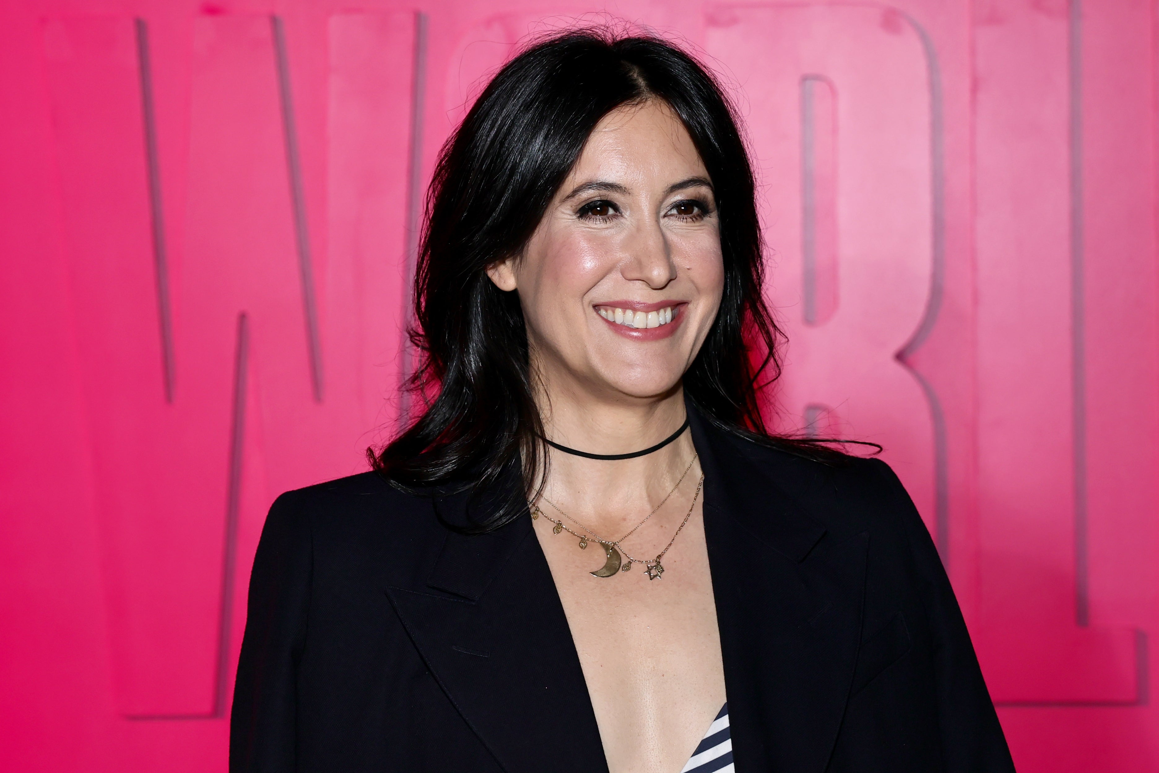 Person smiling, wearing a black blazer and a choker necklace with moon charms, standing in front of a backdrop with large letters