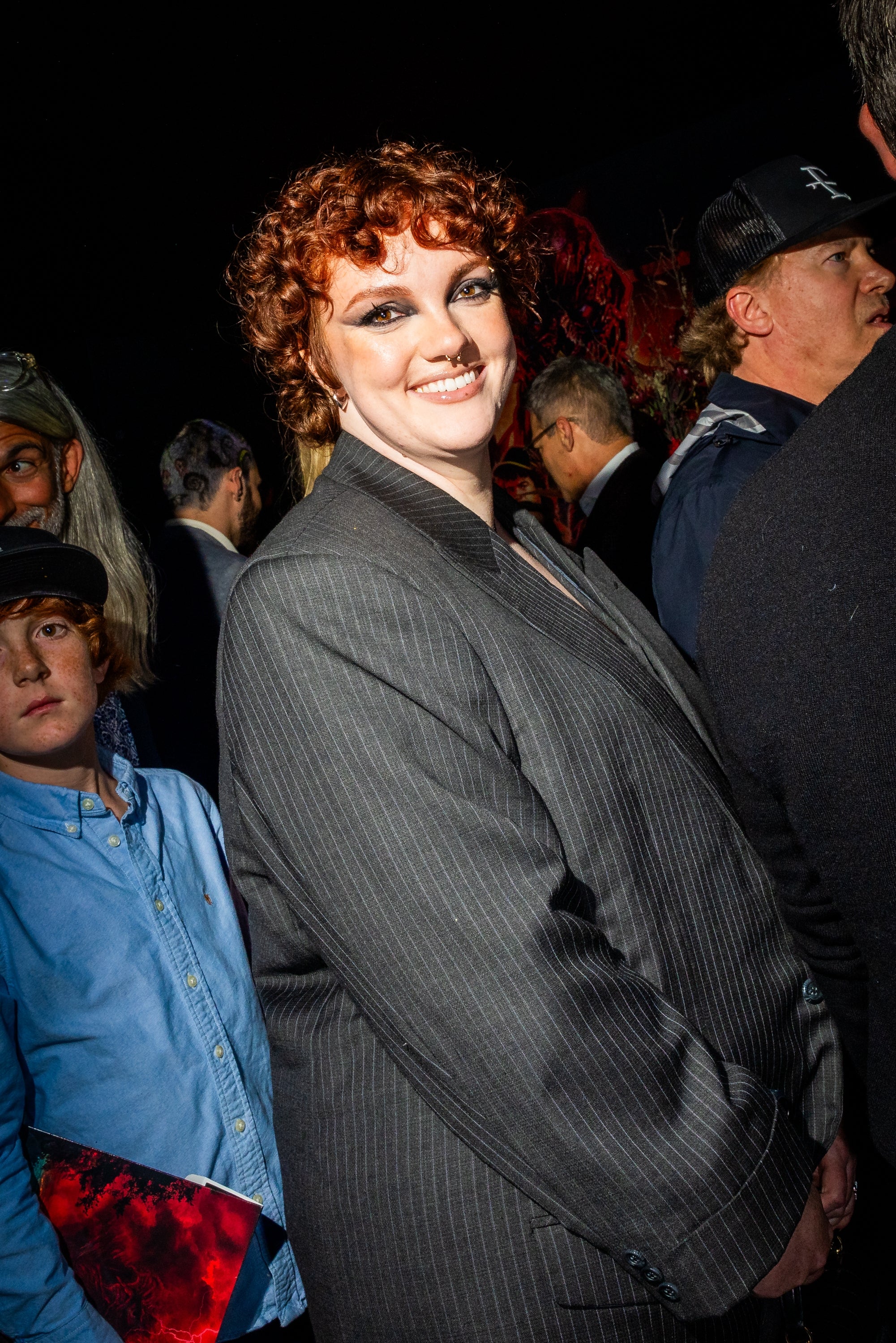 Person with curly hair in a pinstripe suit smiles on a crowded red carpet, standing out in a lively event atmosphere