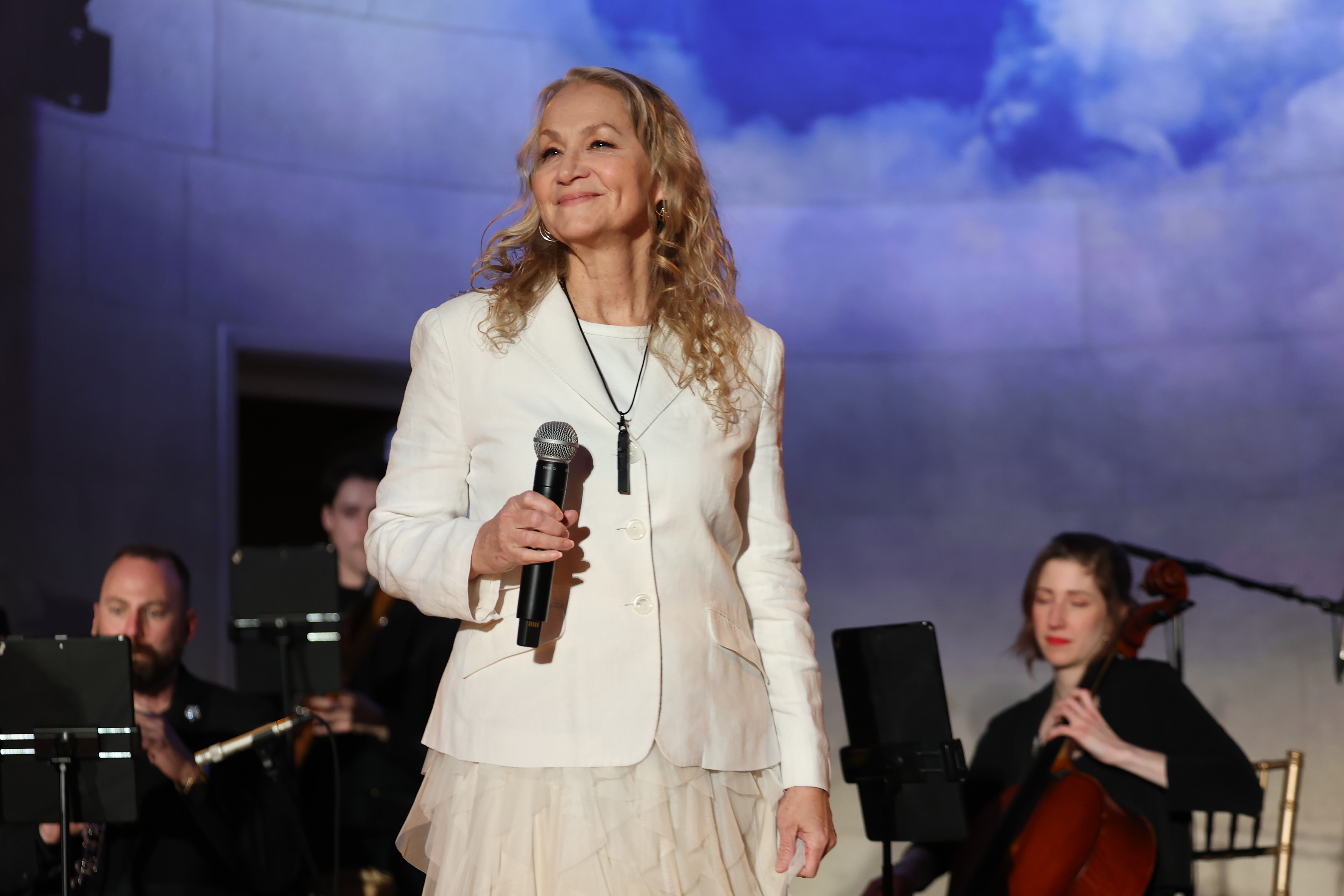 A person wearing a stylish white suit holds a microphone on stage. Background features musicians and a cloud mural