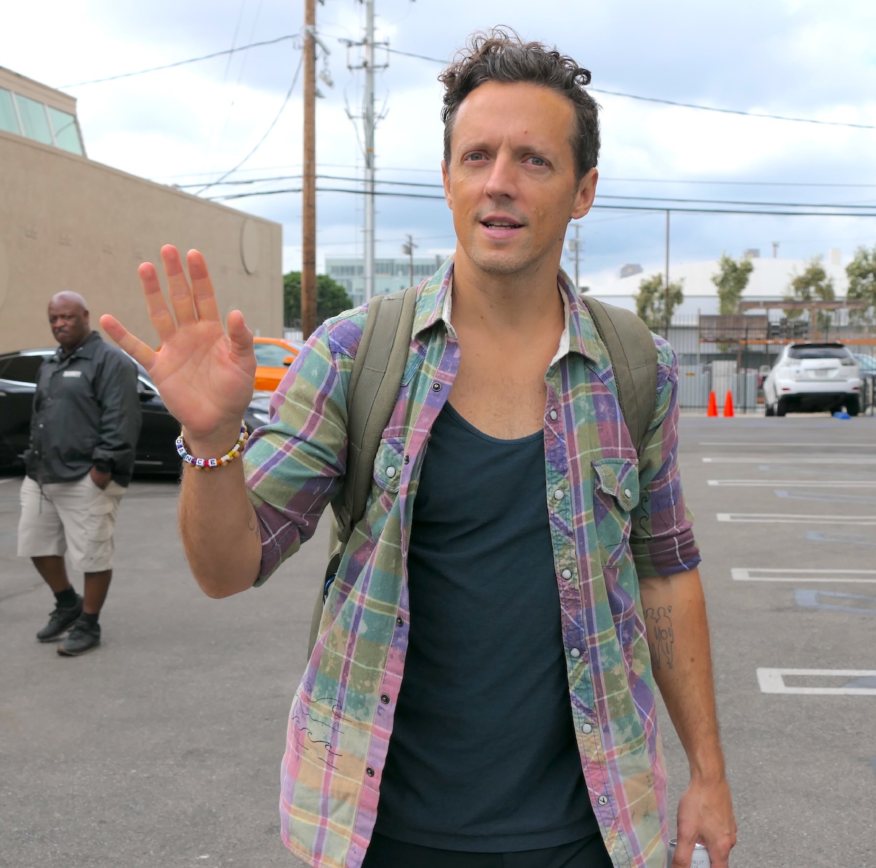 Person wearing a casual plaid shirt, tank top, and backpack, gesturing with one hand in an outdoor setting
