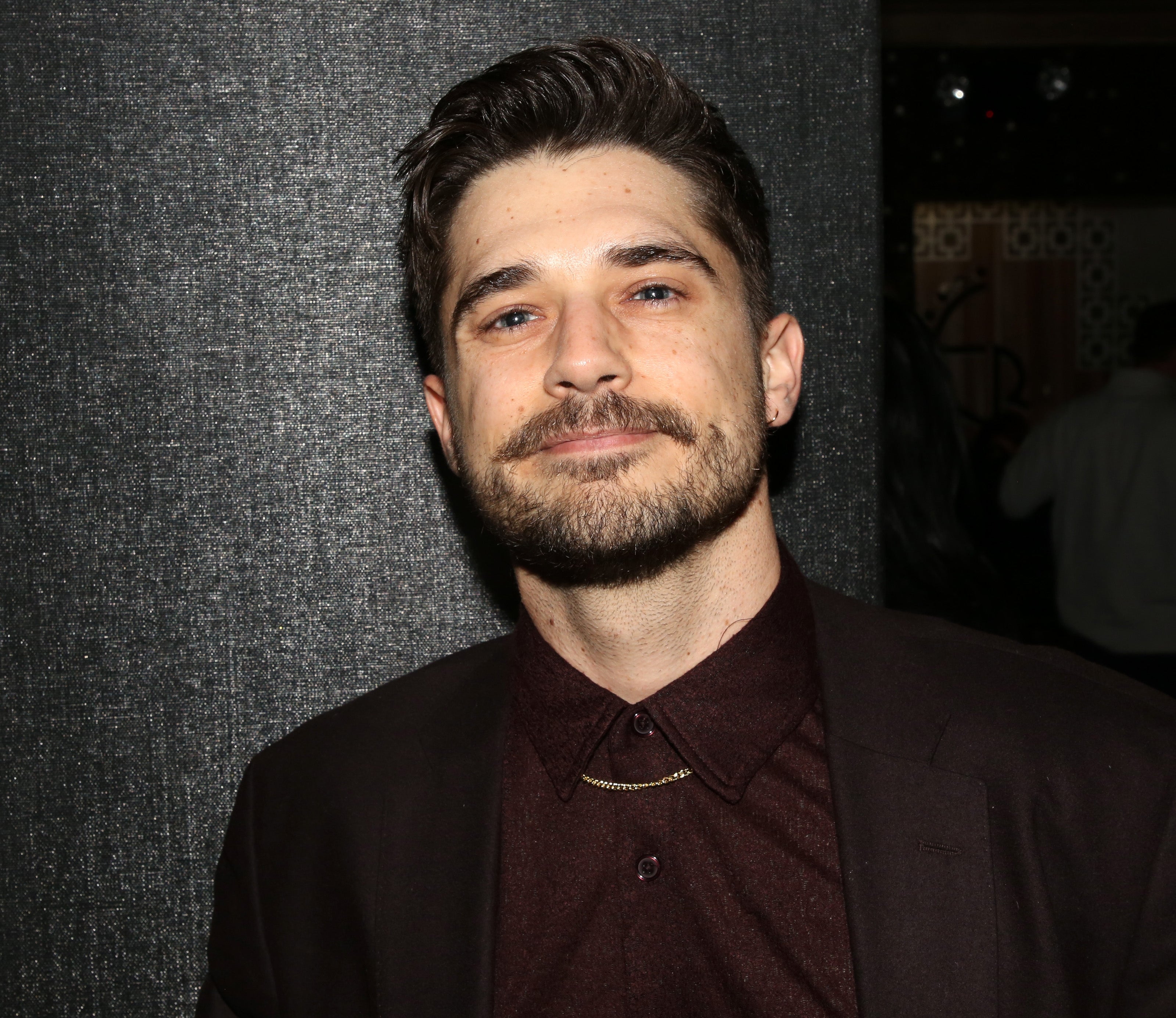 A person with short hair and a beard wears a suit with a collared shirt and chain necklace, posing against a textured background