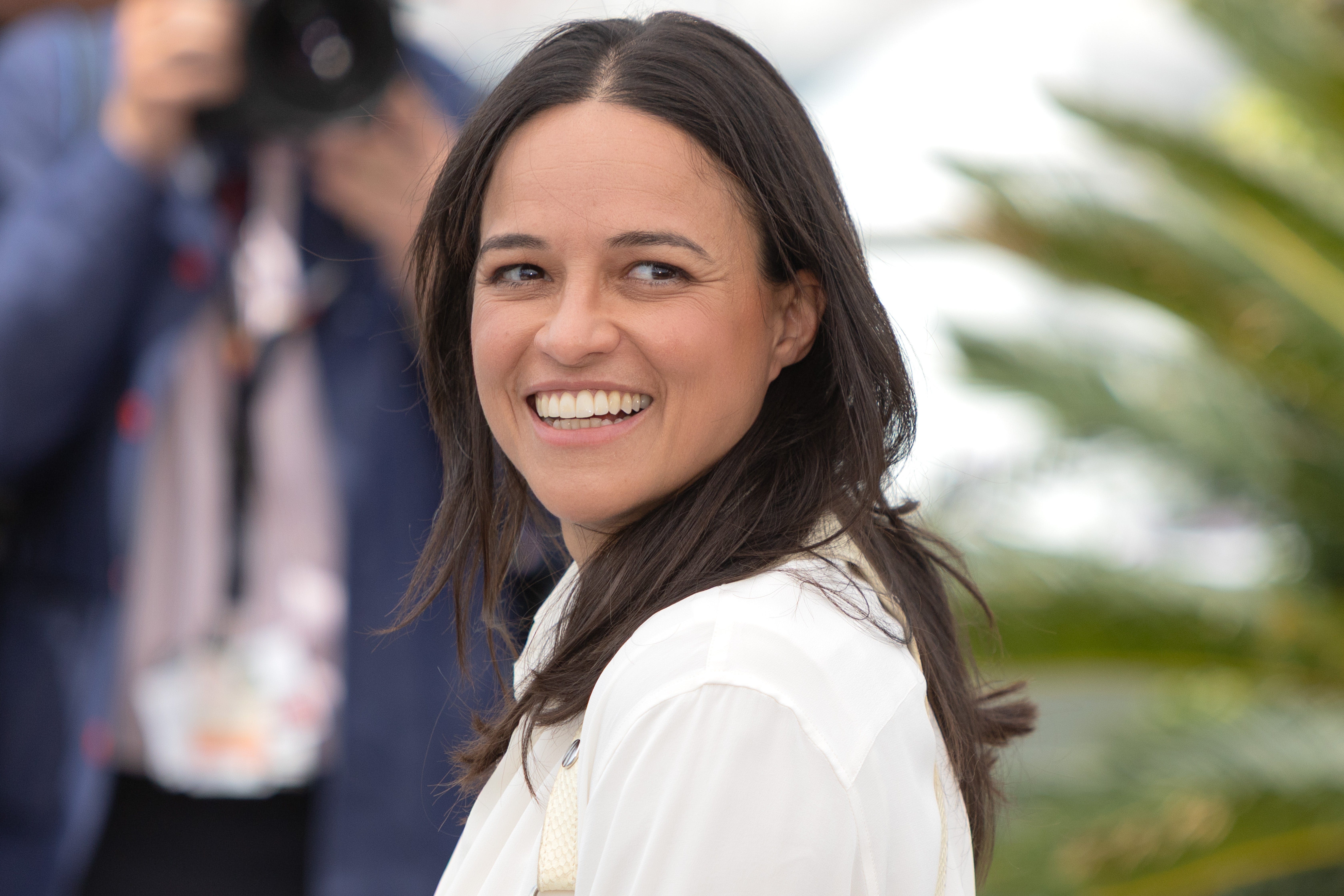 A person smiling at an outdoor event, wearing a white shirt. Photographers are in the background