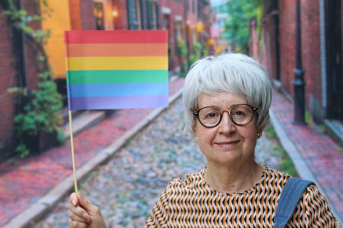 Elderly person with short gray hair holds a rainbow flag, smiling in a cobblestone alley. Wearing a patterned shirt and glasses